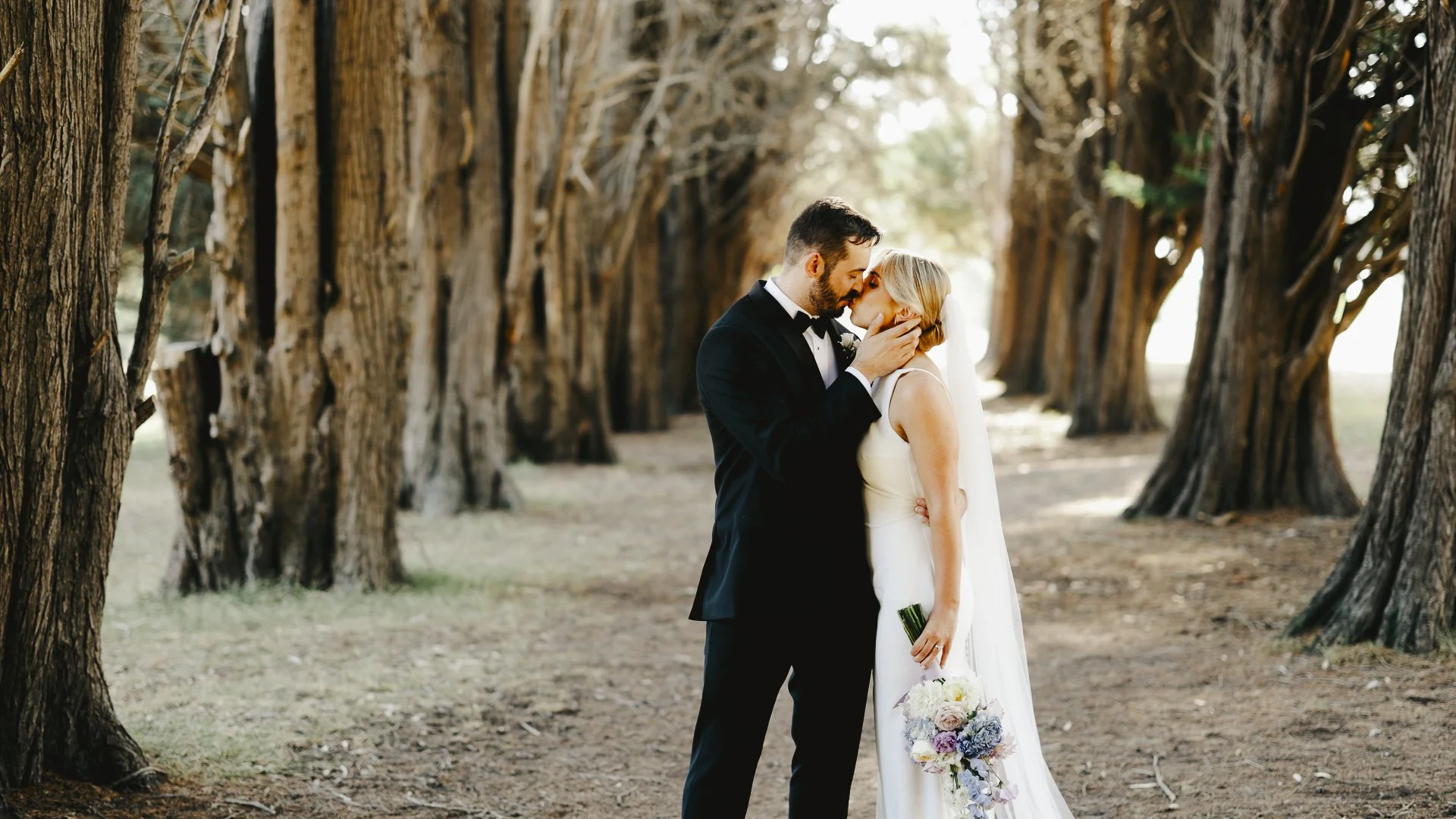 A bride and groom sharing a kiss in a wooded area with tall trees lining the background during their wedding.