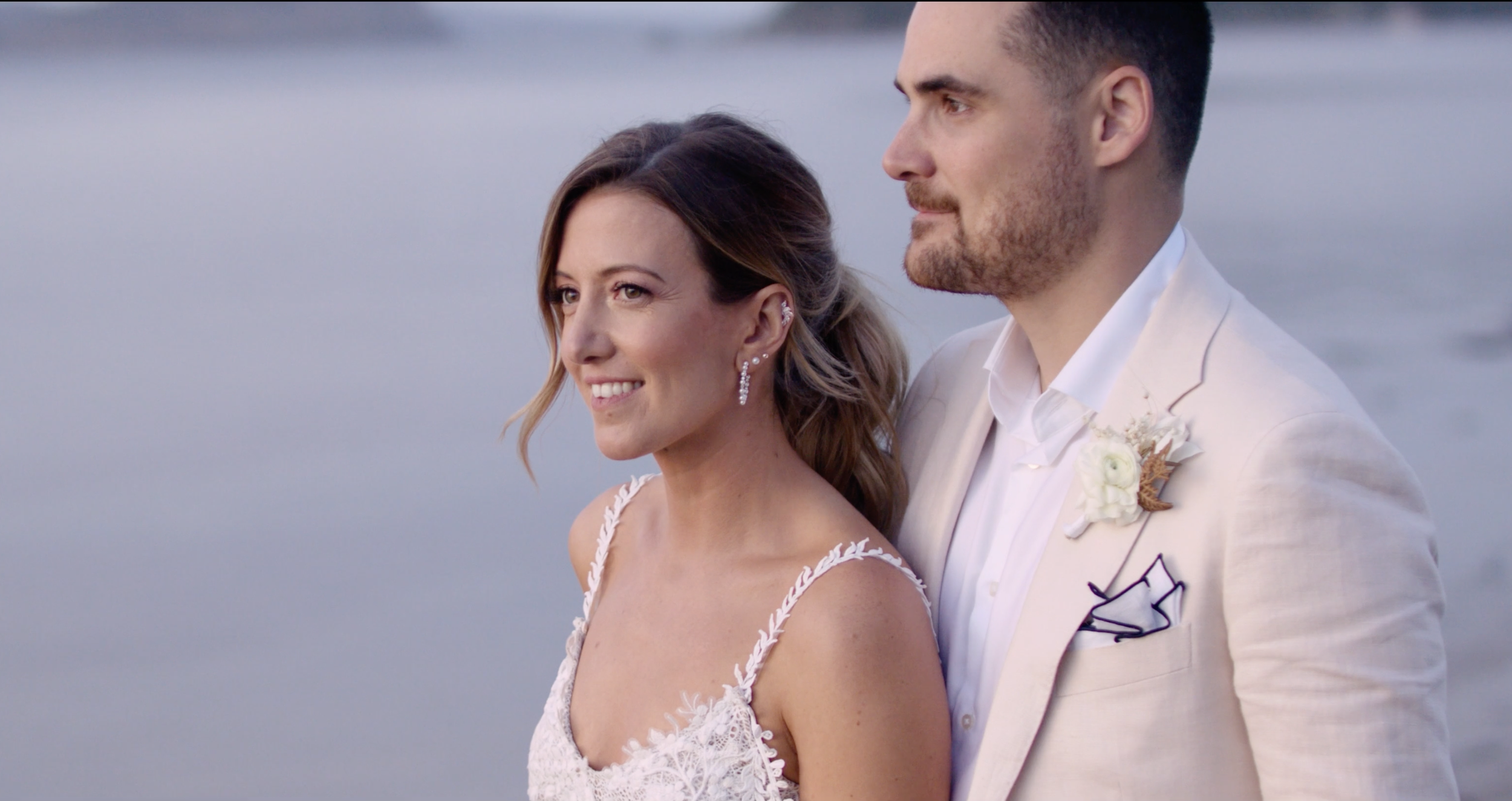 A bride and groom standing by the water during their wedding, with the bride smiling and the groom looking into the distance.