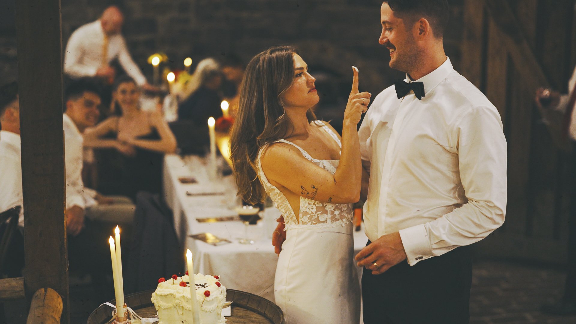 A couple is dancing at a wedding reception, with the woman flicking her finger at the man's face. The woman has long hair, tattoos on her arm, and is wearing a white lace dress. The man has short hair, beard, and is wearing a white shirt and black bow tie. There is a wedding cake with candles on a table nearby, and guests are seated at tables in the background, with warm lighting and a rustic setting.