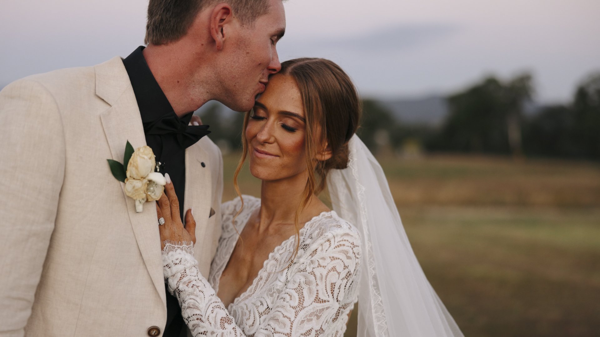 A bride and groom on their wedding day. The groom is wearing a cream-colored suit with a black shirt and bow tie, and the bride is dressed in a lace wedding gown with a veil. The groom is kissing the bride on the forehead, and she is smiling with her eyes closed. They are outdoors with a blurred landscape in the background.