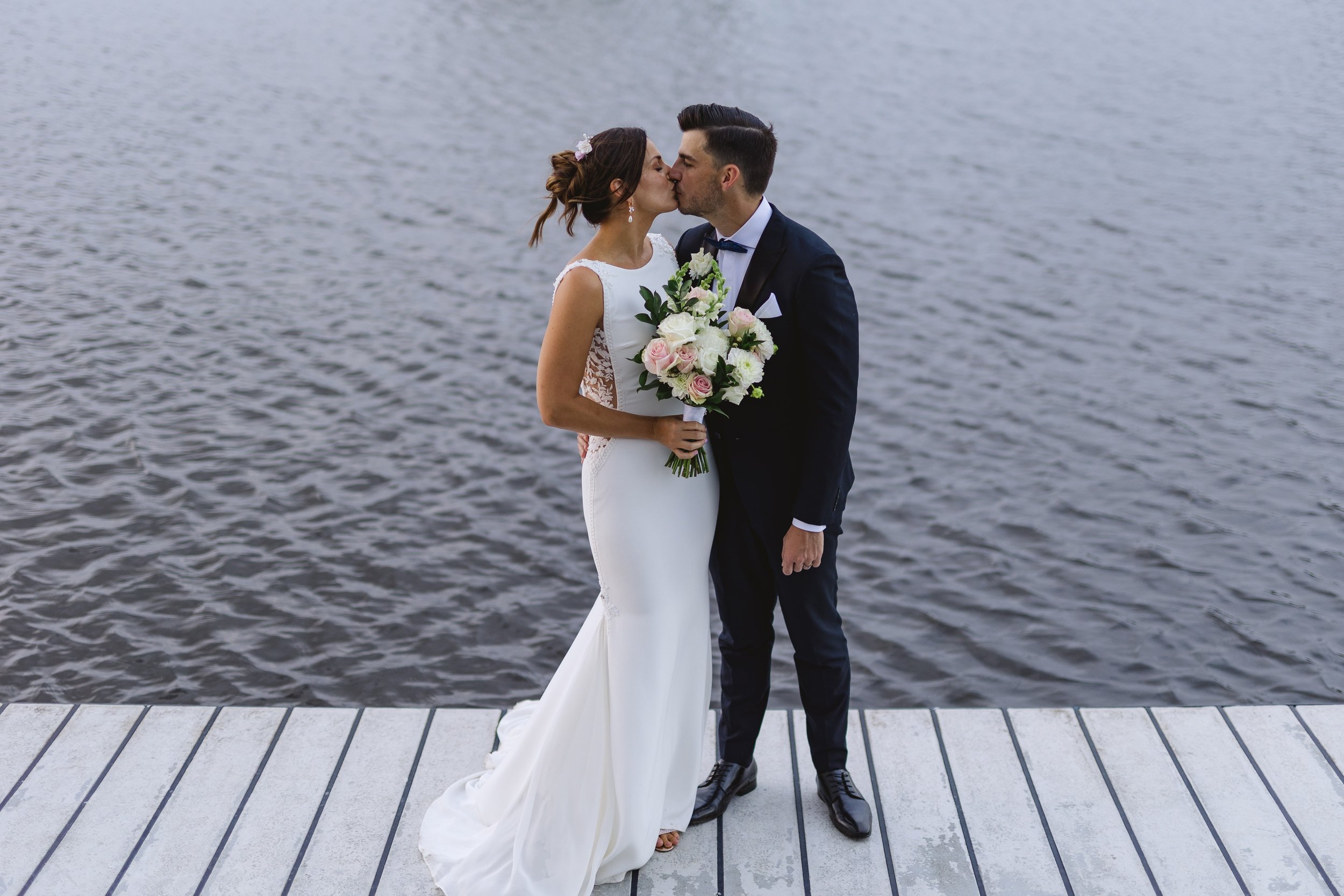 A bride and groom kissing by a body of water on a dock, with the bride holding a bouquet of pink and white roses and greenery, both dressed in wedding attire.