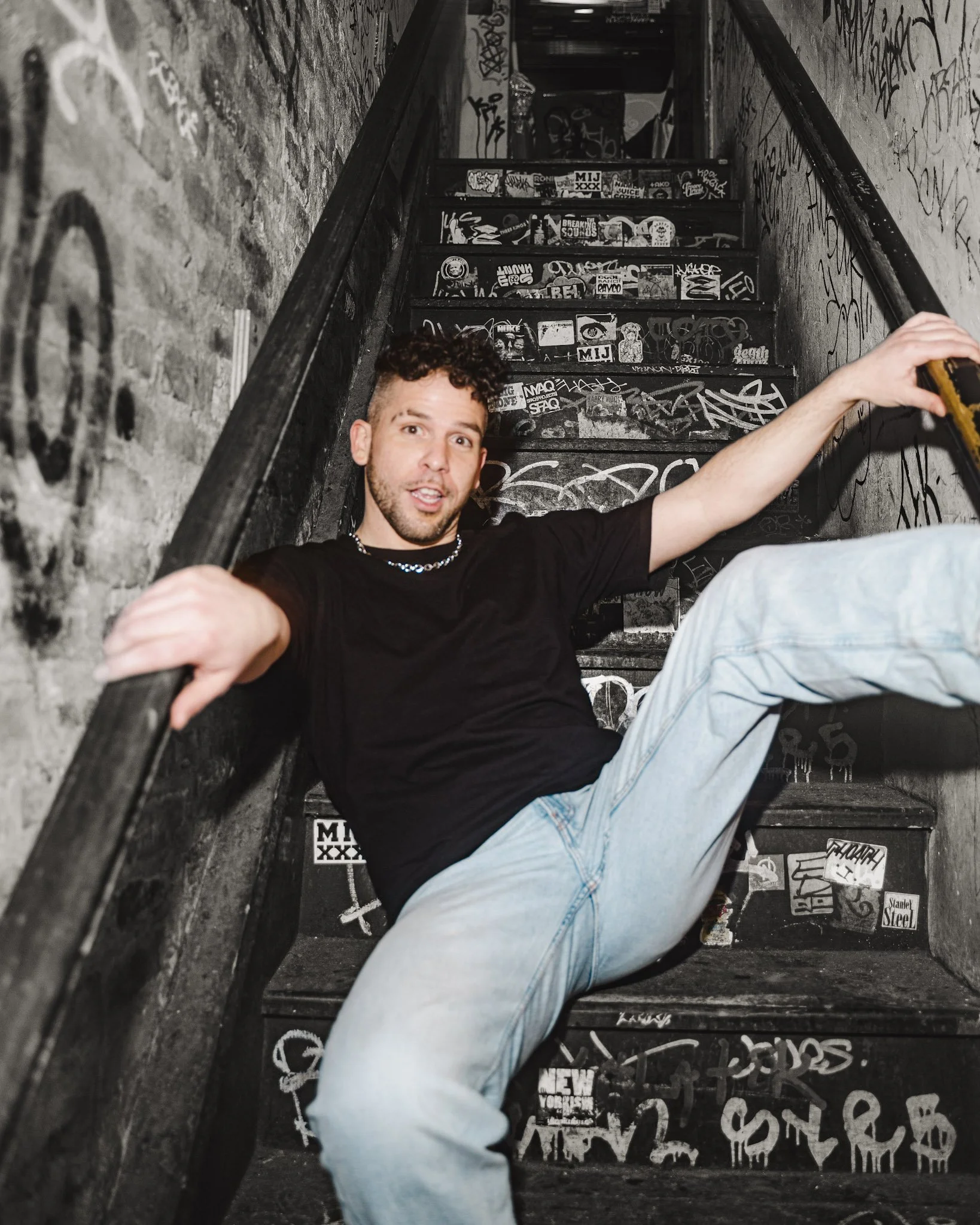 Steven Zakari sitting on graffiti-covered stairs in a dimly lit alley, holding onto the railing with one arm extended, wearing a black t-shirt and light jeans.