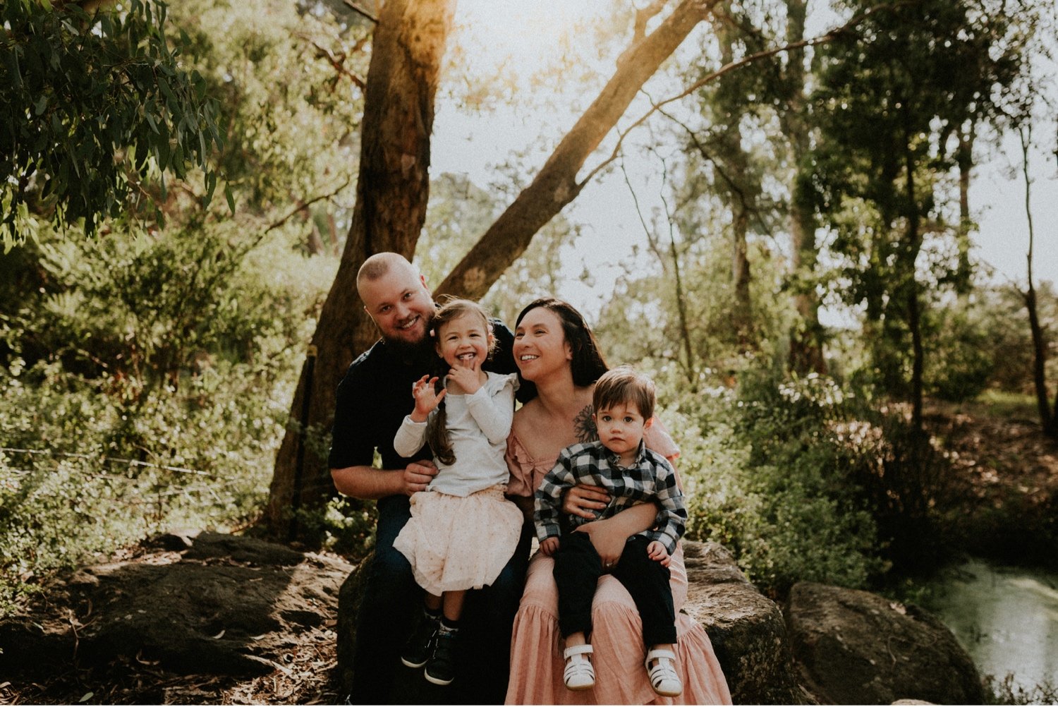 18_DTEV-79_Family portrait sitting on a rock with the sun shining behind them, piercing through the trees. .jpg