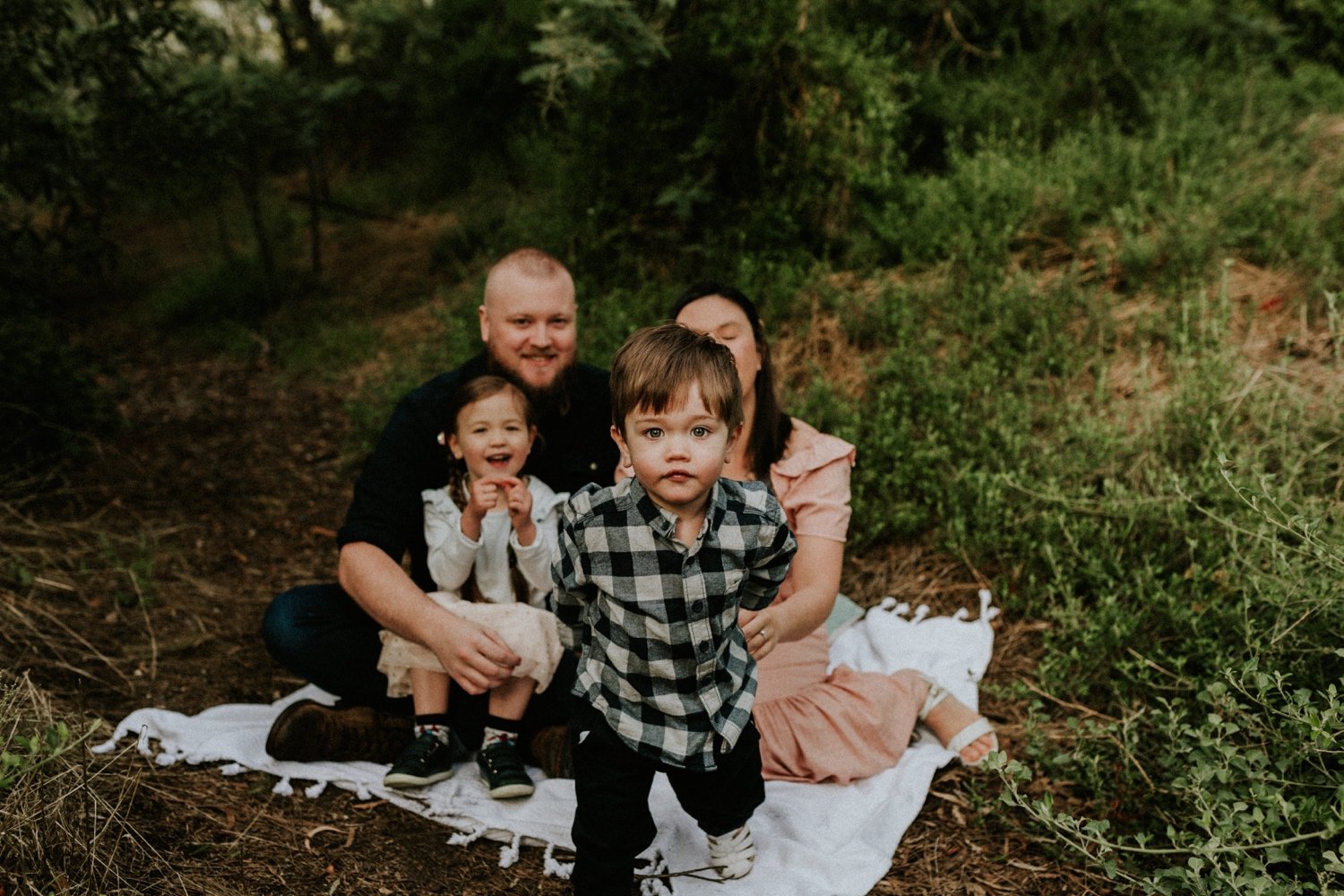 04_DTEV-14_2 year old boy running towards the camera in the middle of a family session, while mum, dad and sister sit behind him, look at him and smile. .jpg