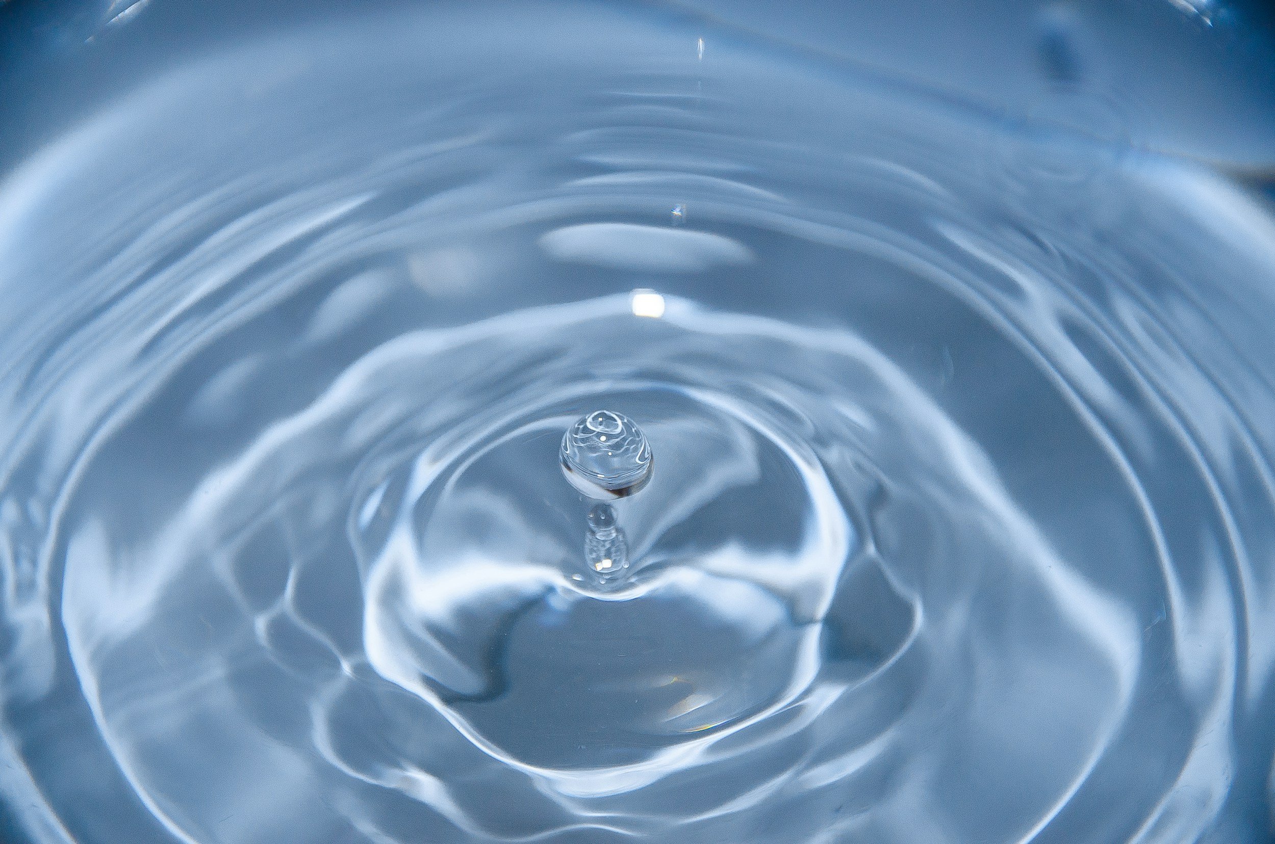 Close-up image of a single drop of water rising from the surface of a calm pool, creating concentric ripples that spread outward in soft blue tones. The water is smooth and reflective, capturing a sense of fluidity, stillness, and gentle movement.