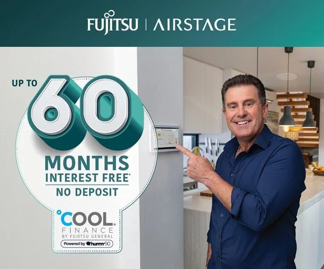 Smiling man in a blue shirt pointing to a promotional sign for a financing offer, with a kitchen in the background. The sign advertises up to 60 months interest free with no deposit from Fujitsu Airstage and Cool Finance.