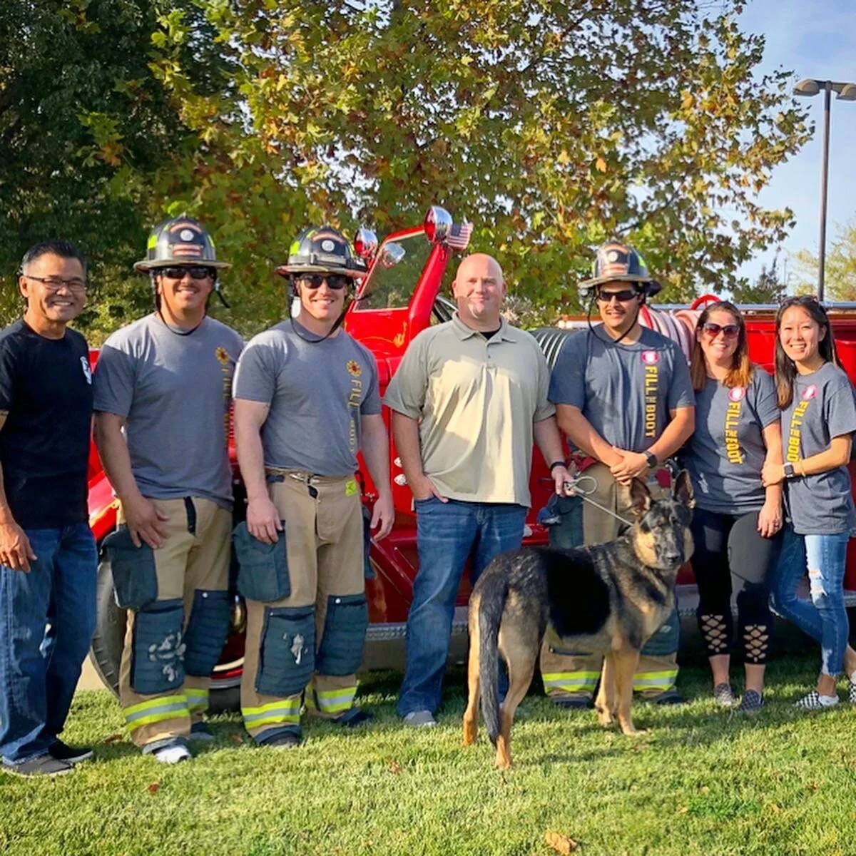 Group of firefighters and civilians standing in front of a red fire truck, with some firefighters wearing helmets and firefighting gear, and a German Shepherd dog on a leash.