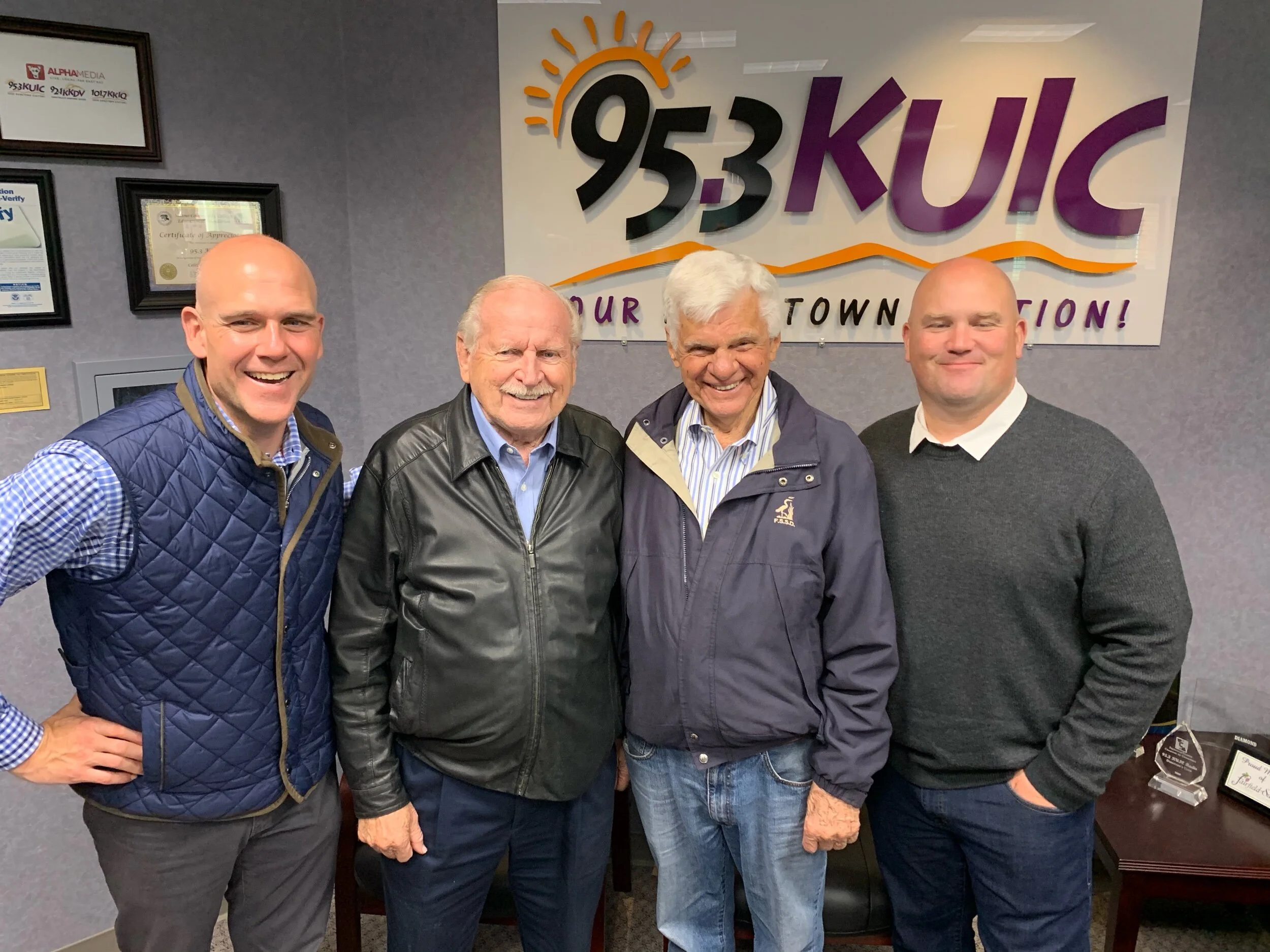 Four men standing together in front of a wall with a sign that reads '95.3 KUIC, Your Town Station!' and framed certificates. The men are smiling and appear happy.