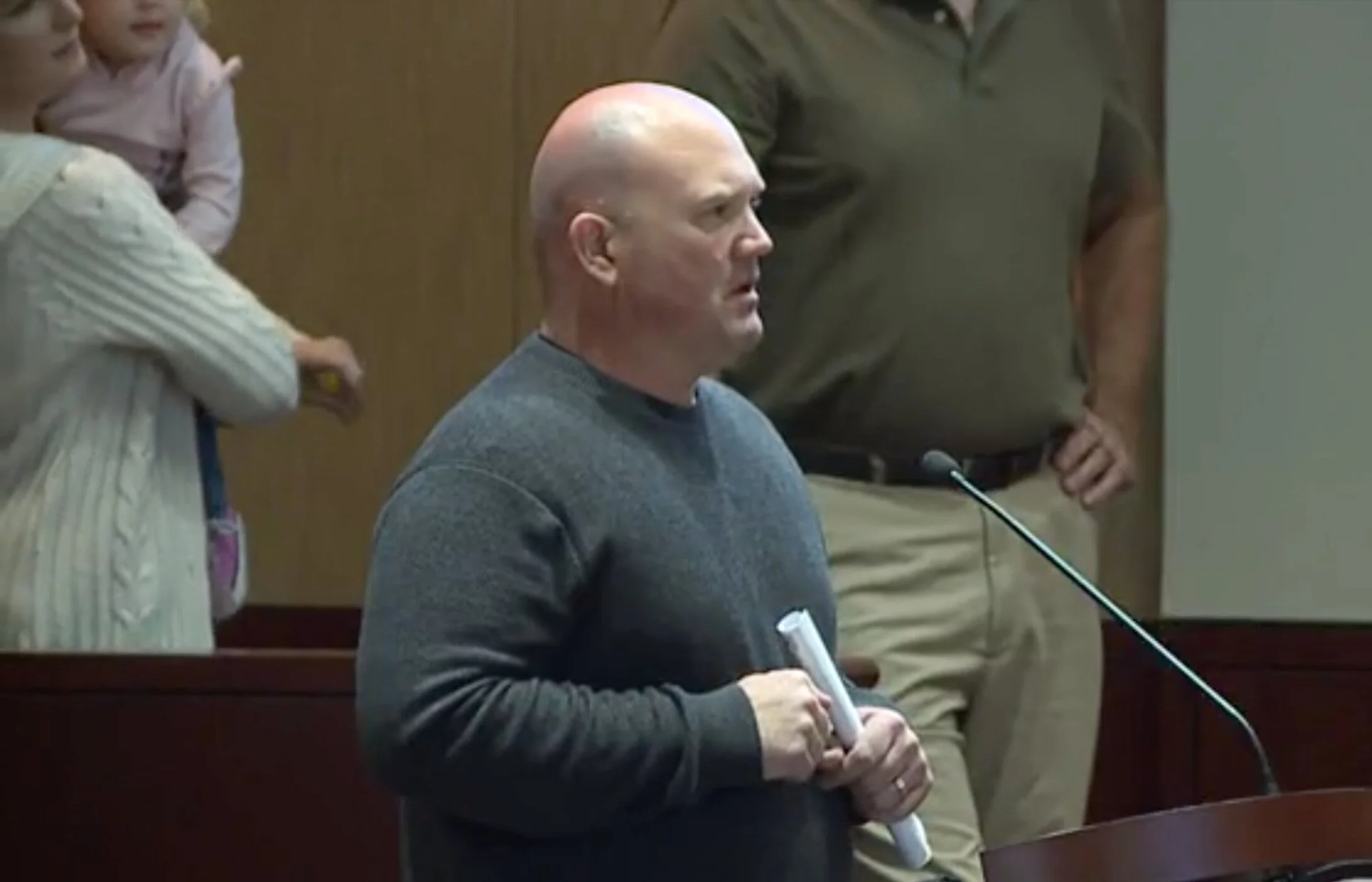 A man with a bald head wearing a gray sweater is sitting at a table with a pen and paper. He appears to be at a hearing or conference, with microphones and other people standing around him.