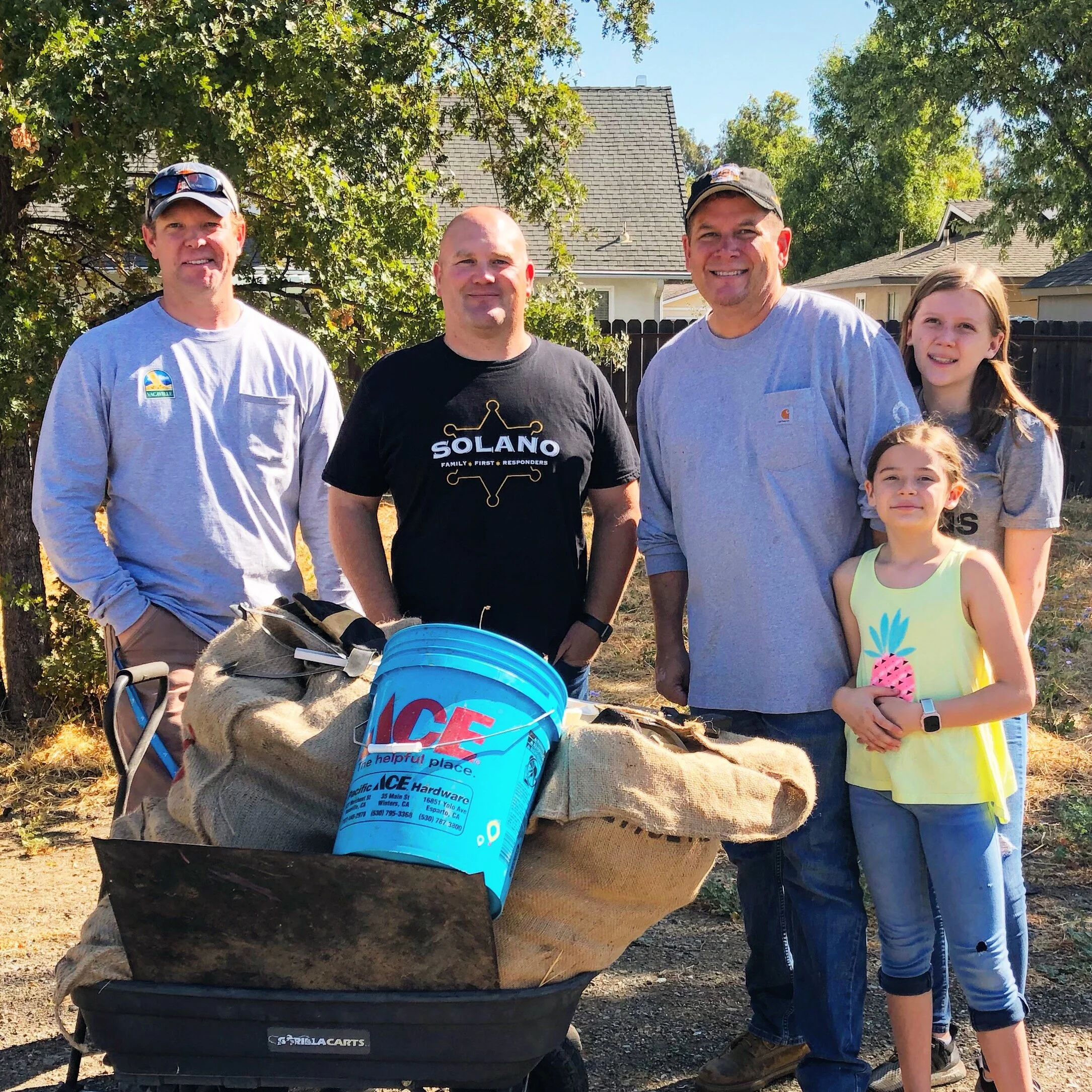 Four people, two men and two young girls, are standing behind a large collection of bags and a blue ice chest, outdoors in front of a house with trees. They are smiling and dressed casually, with the men wearing caps and T-shirts, and the girls wearing tank tops.