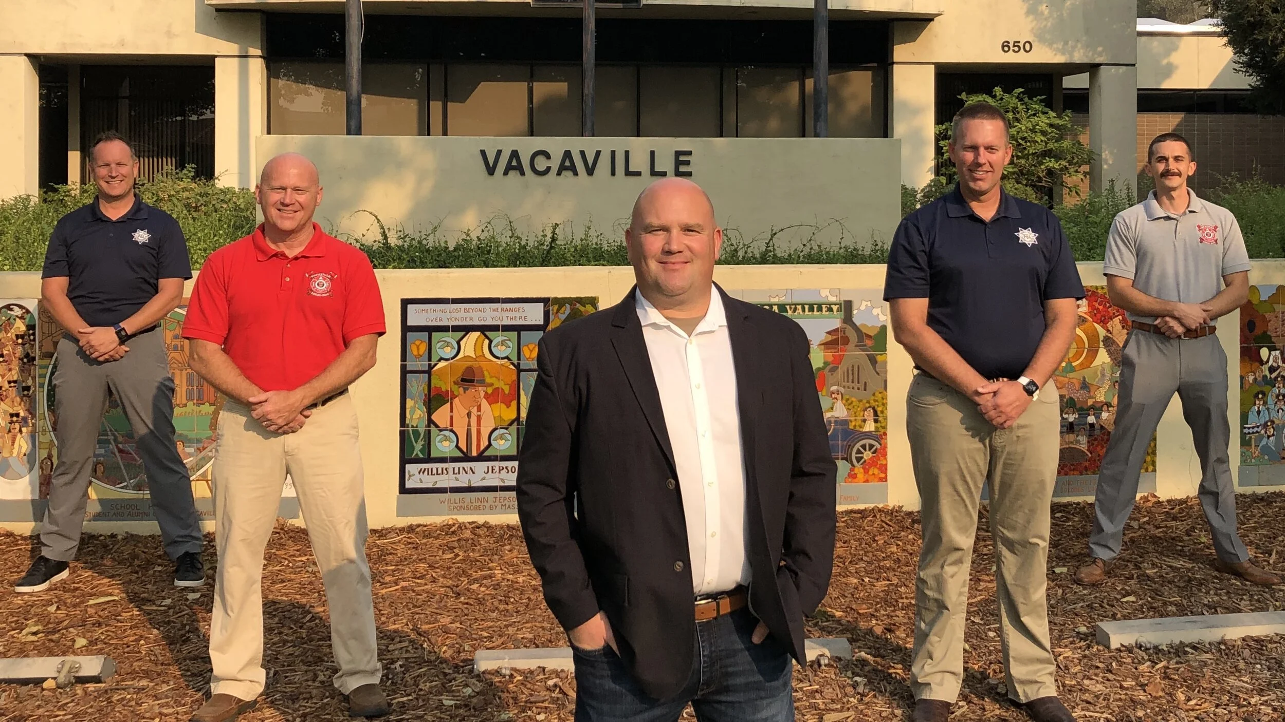 Six men standing outdoors in front of a building with a sign that reads 'Vacaville.' They are dressed in business or casual attire, with some wearing polo shirts featuring badges or logos. The group is positioned on mulch and there are colorful mosaic art pieces behind them. One man in a black suit stands in the foreground, smiling at the camera.