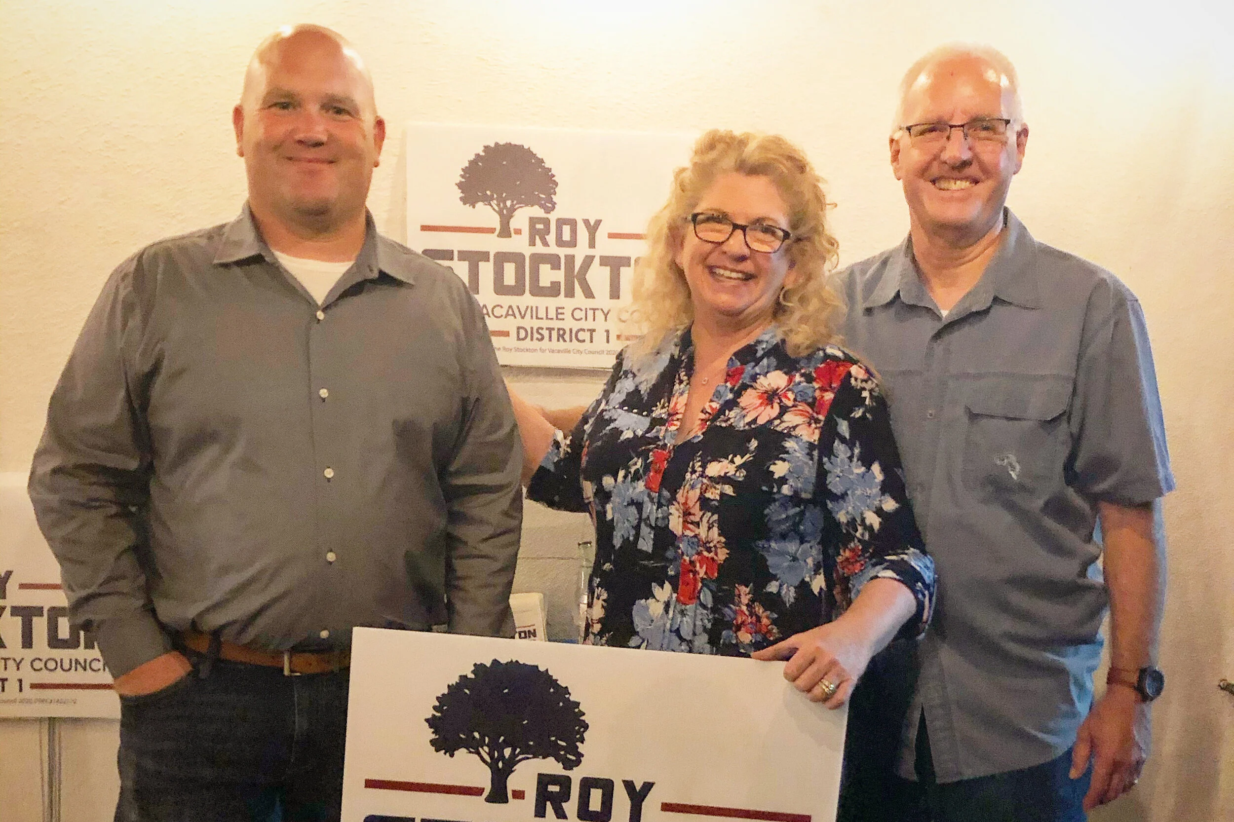 Three people standing together, smiling, at a political event, with a sign showing a tree logo and the text 'ROY STOCKTON' in the background.