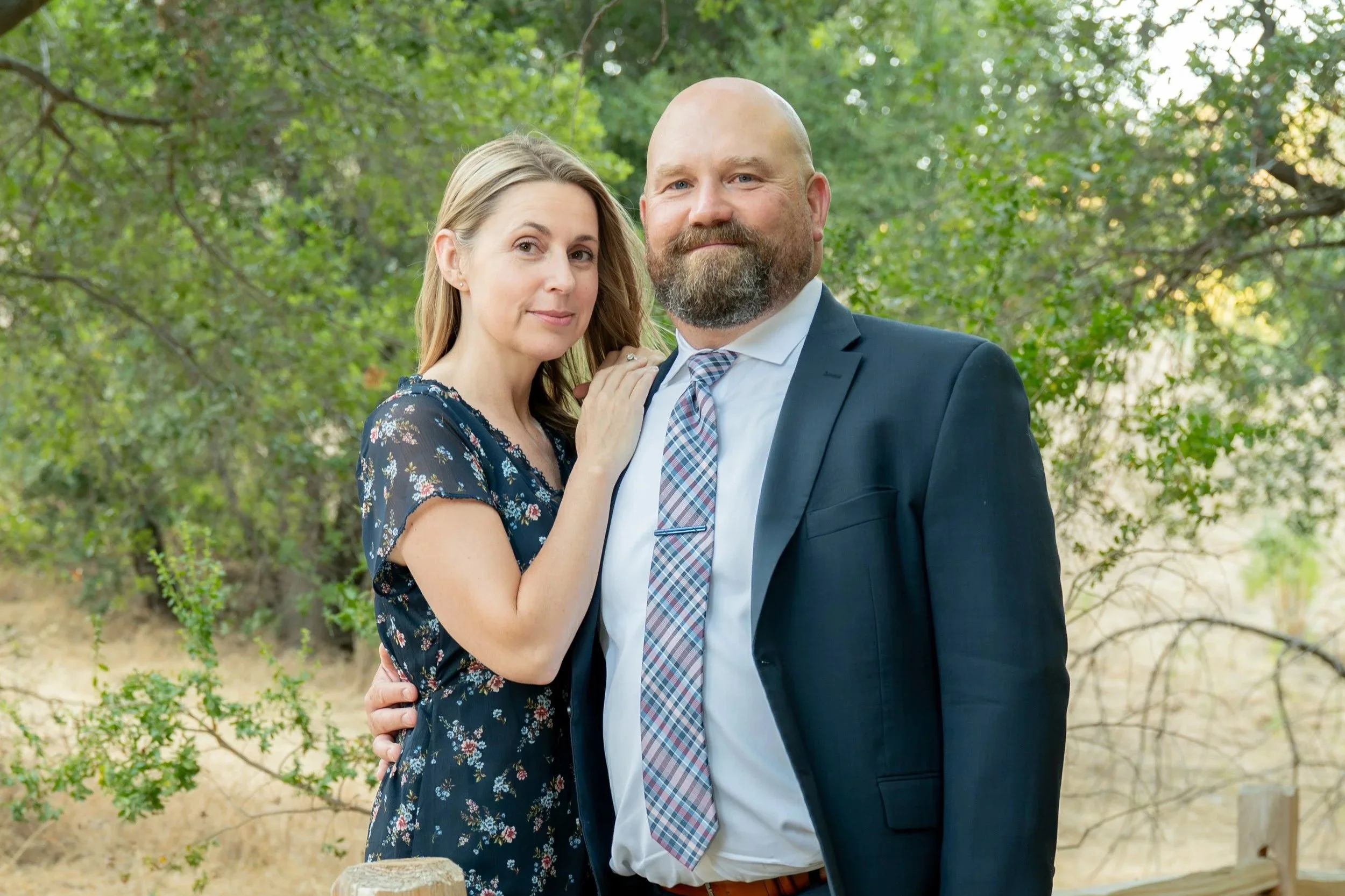 A couple posing outdoors in a wooded area, the woman is wearing a floral dress and the man is in a suit with a tie.