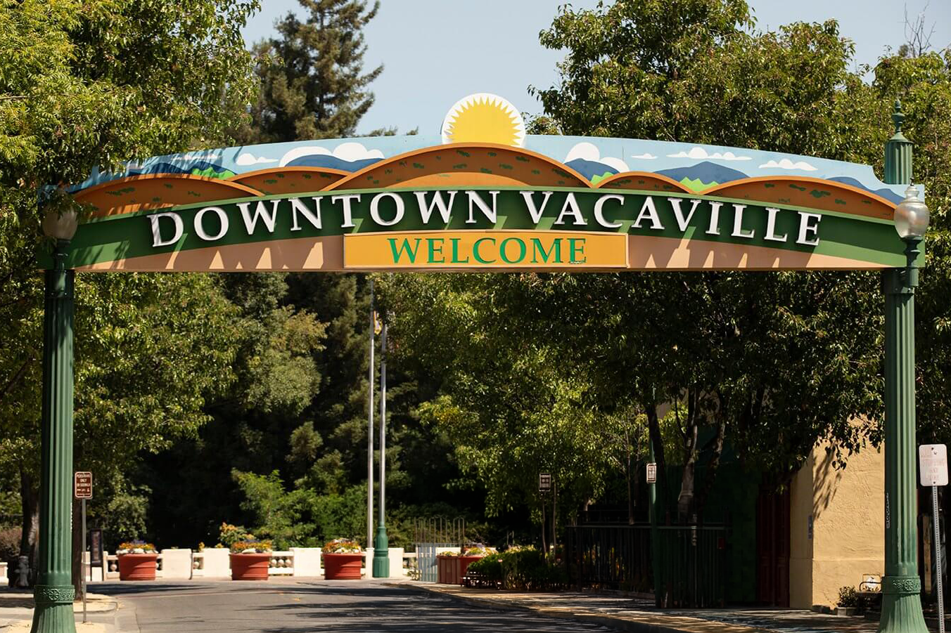Sign reading 'Downtown Vacaville Welcome' above a street entrance, with trees and buildings in the background.