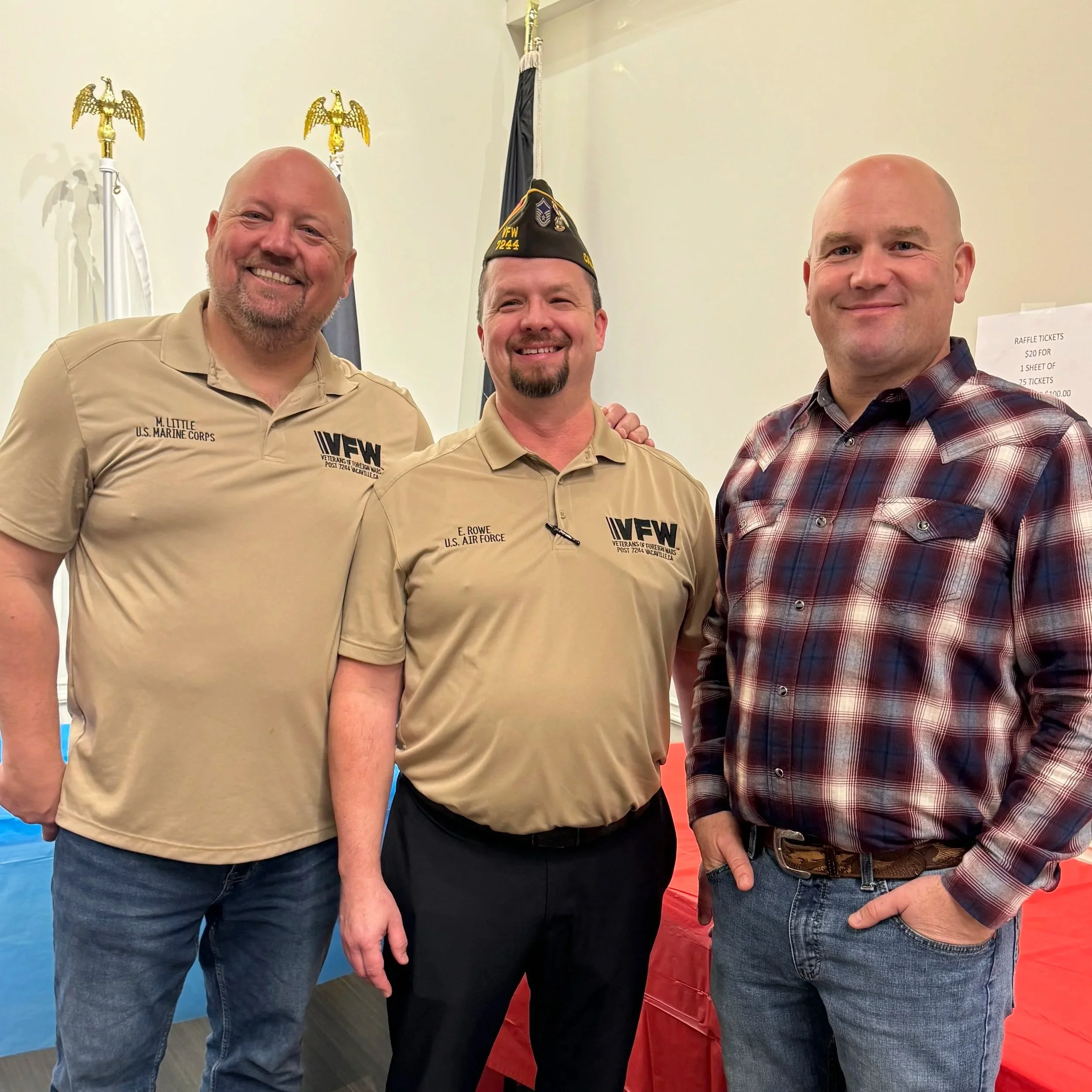 Three men smiling at the camera, two of them are wearing beige U.S. Marine Corps shirts with one also wearing a veterans of foreign wars baseball cap, the third man is wearing a plaid shirt, possible at a veterans or military event with American flags and patriotic decorations in the background.