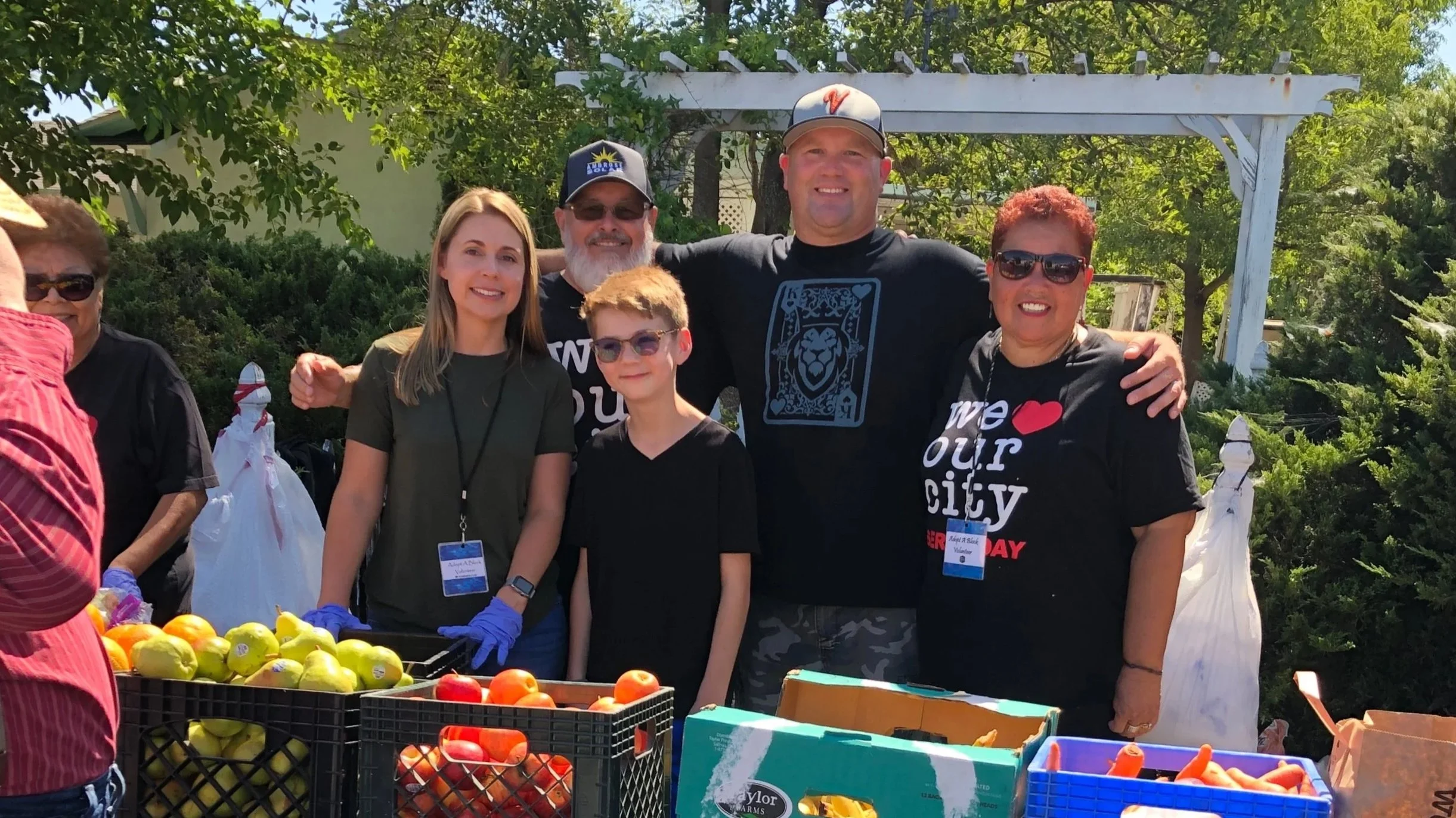 Group of six smiling people at a farmers market, standing behind crates of apples and peaches, with trees and a white trellis in the background.