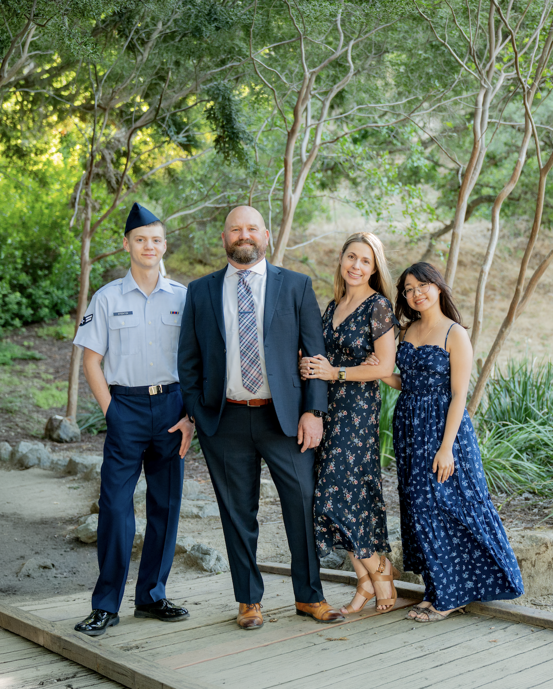 A family of four standing outdoors on a wooden platform amidst trees and greenery, posing for a photo. The family includes a young man in a military uniform, a middle-aged man in a suit, a woman in a floral dress, and a young woman in a blue patterned dress.