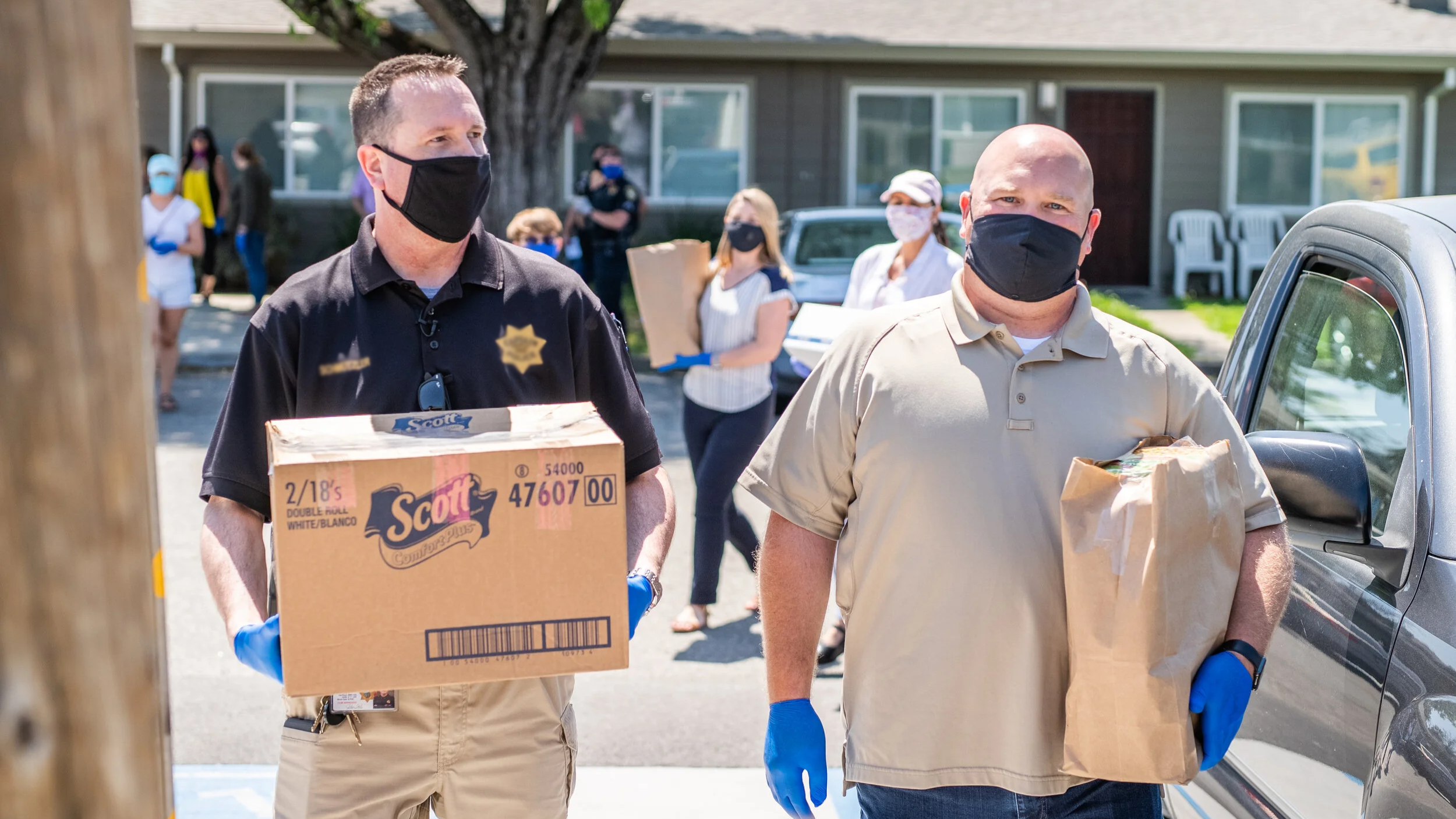 Two men wearing black masks and beige gloves, carrying boxes of supplies, with several people in the background, outdoors in a residential area.