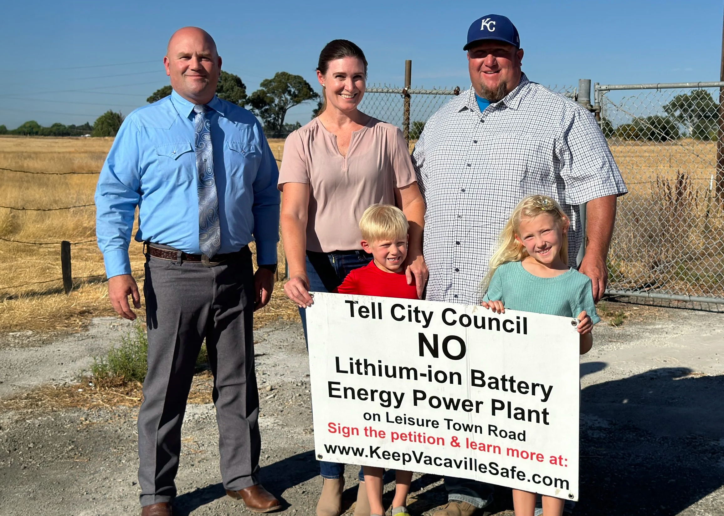 Group of four adults and two children standing outdoors behind a sign protesting lithium-ion battery plant. The adults are dressed in business casual and semi-casual attire, the children are smiling. The background features a fence, open field, and clear blue sky.