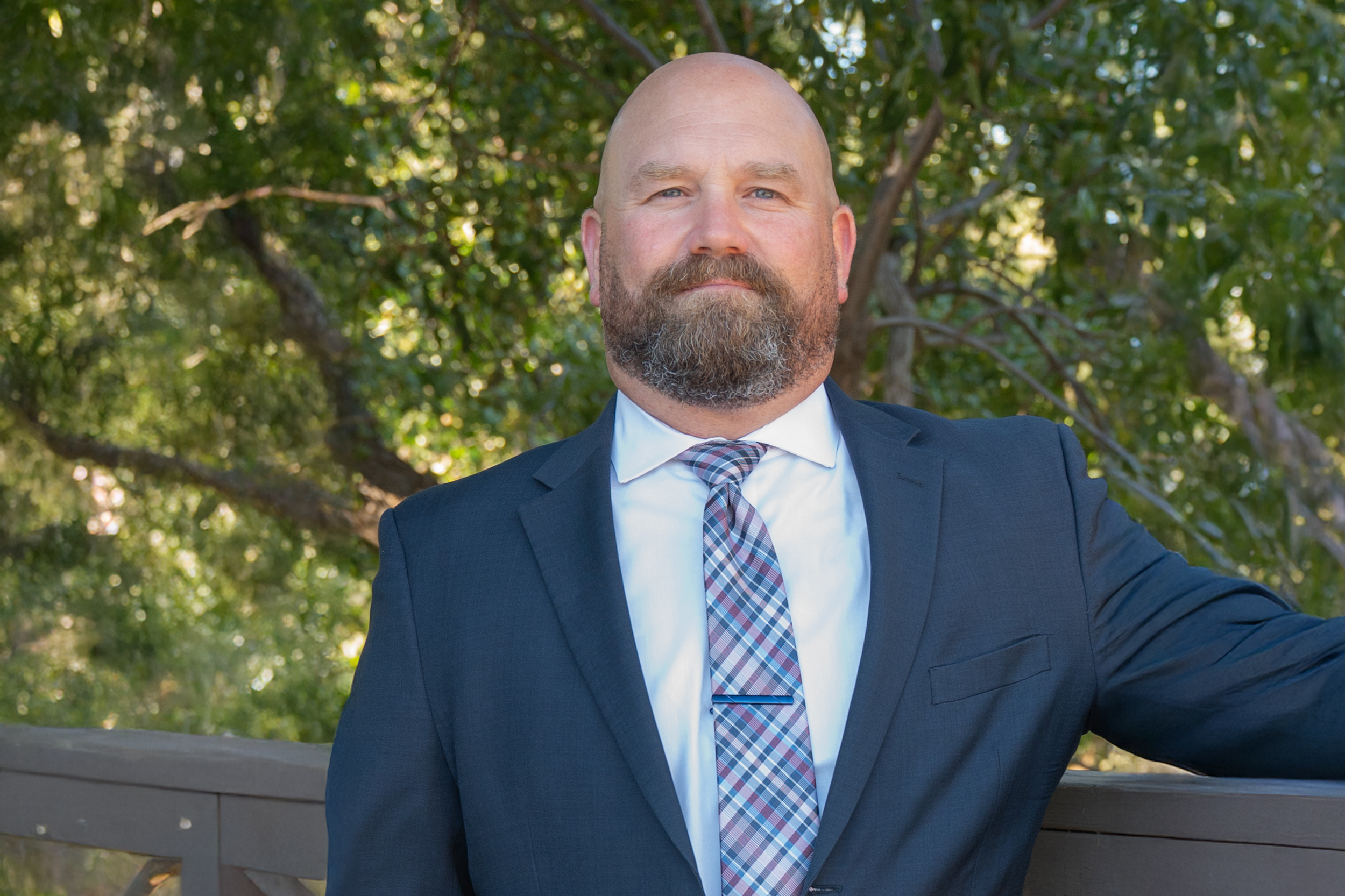 Bald man with a beard wearing a dark suit, white shirt, and plaid tie, standing outdoors in front of green trees.