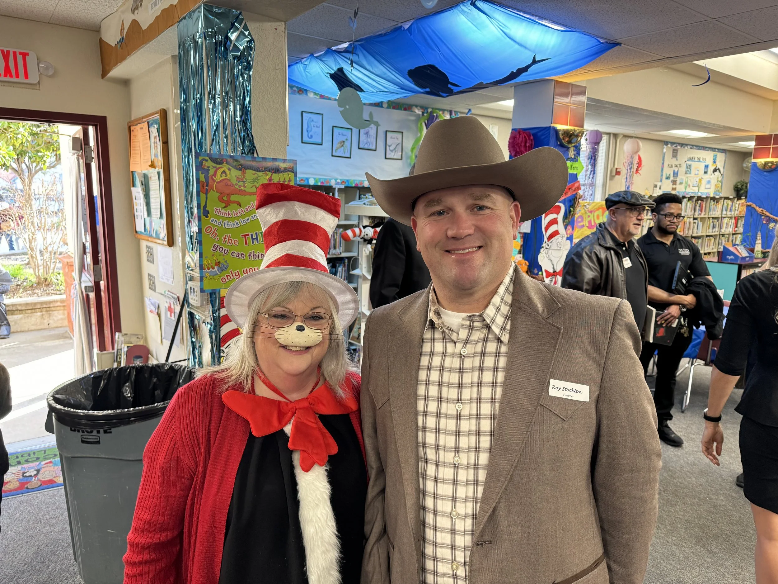 A man in a cowboy hat and beige blazer standing next to a woman in a red cardigan and cat costume at a library decorated with blue and sea-themed decorations.