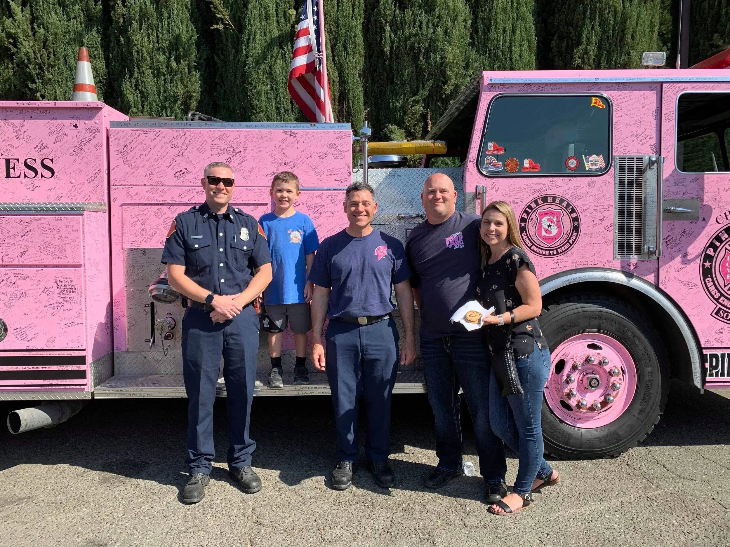 Group of five people standing in front of a pink fire truck covered with signatures and messages, with an American flag in the background.