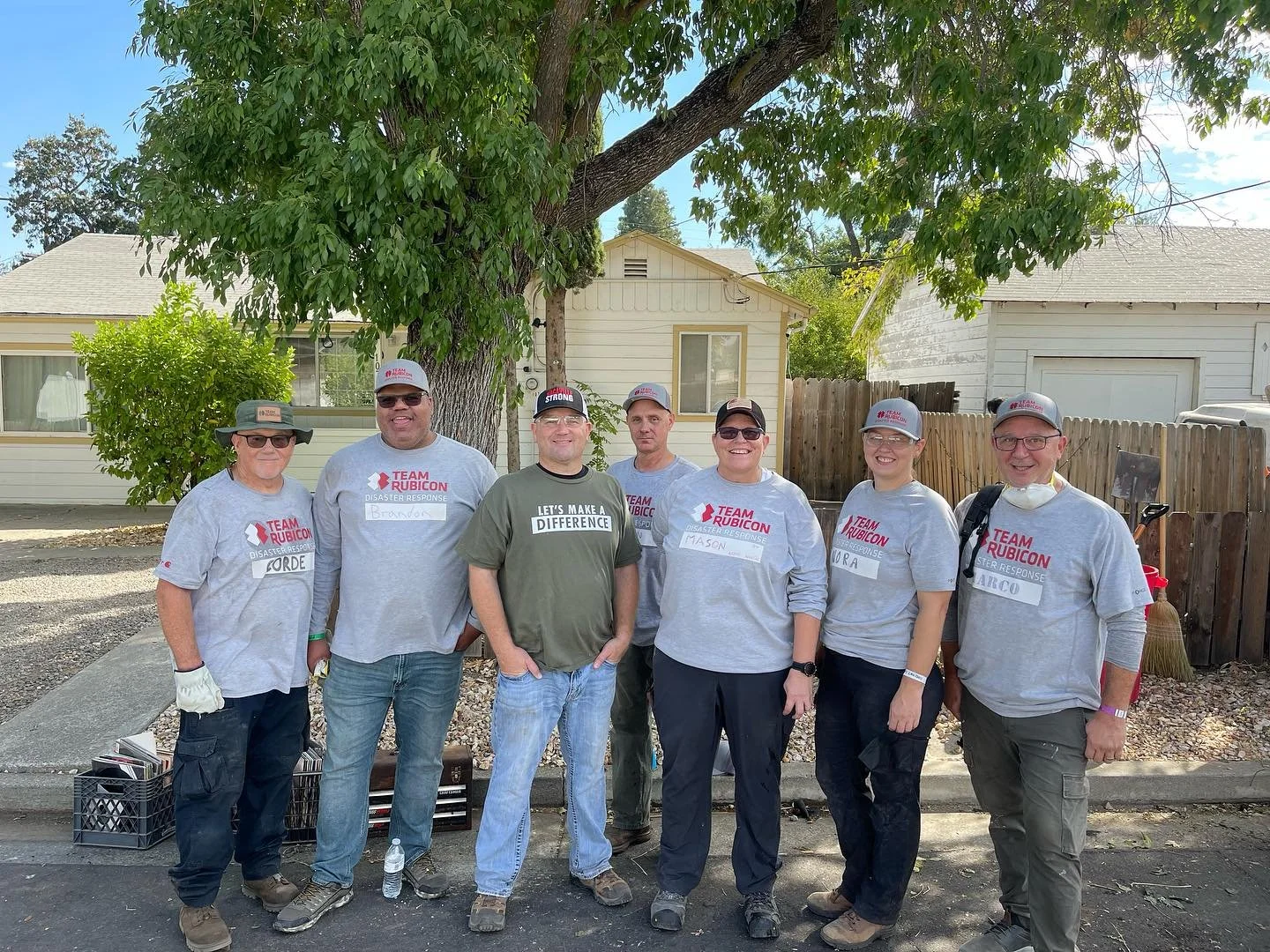 Group of eight people standing outdoors in front of a tree and houses, all wearing gray shirts and hats with team logos, participating in a disaster response event.