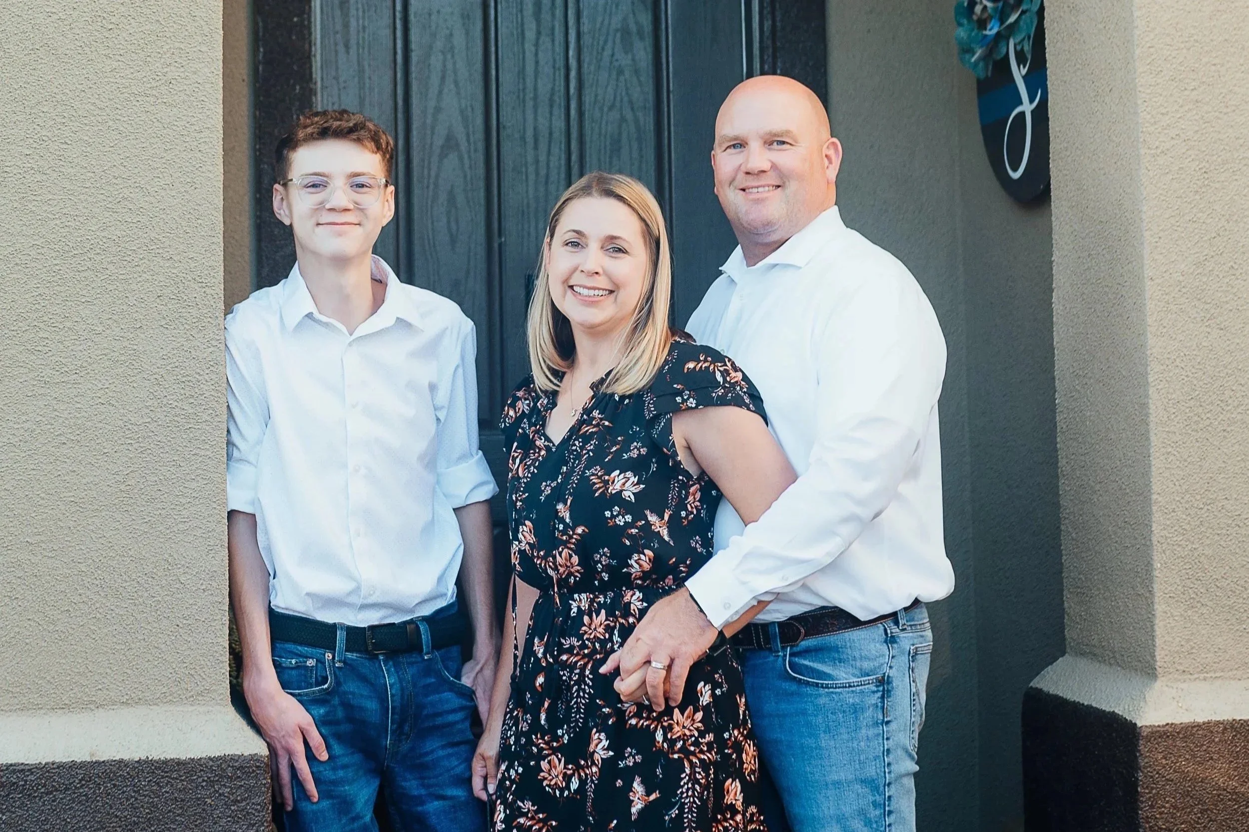 A family of three standing outside a house, smiling at the camera, with a dark door behind them and beige walls on either side.