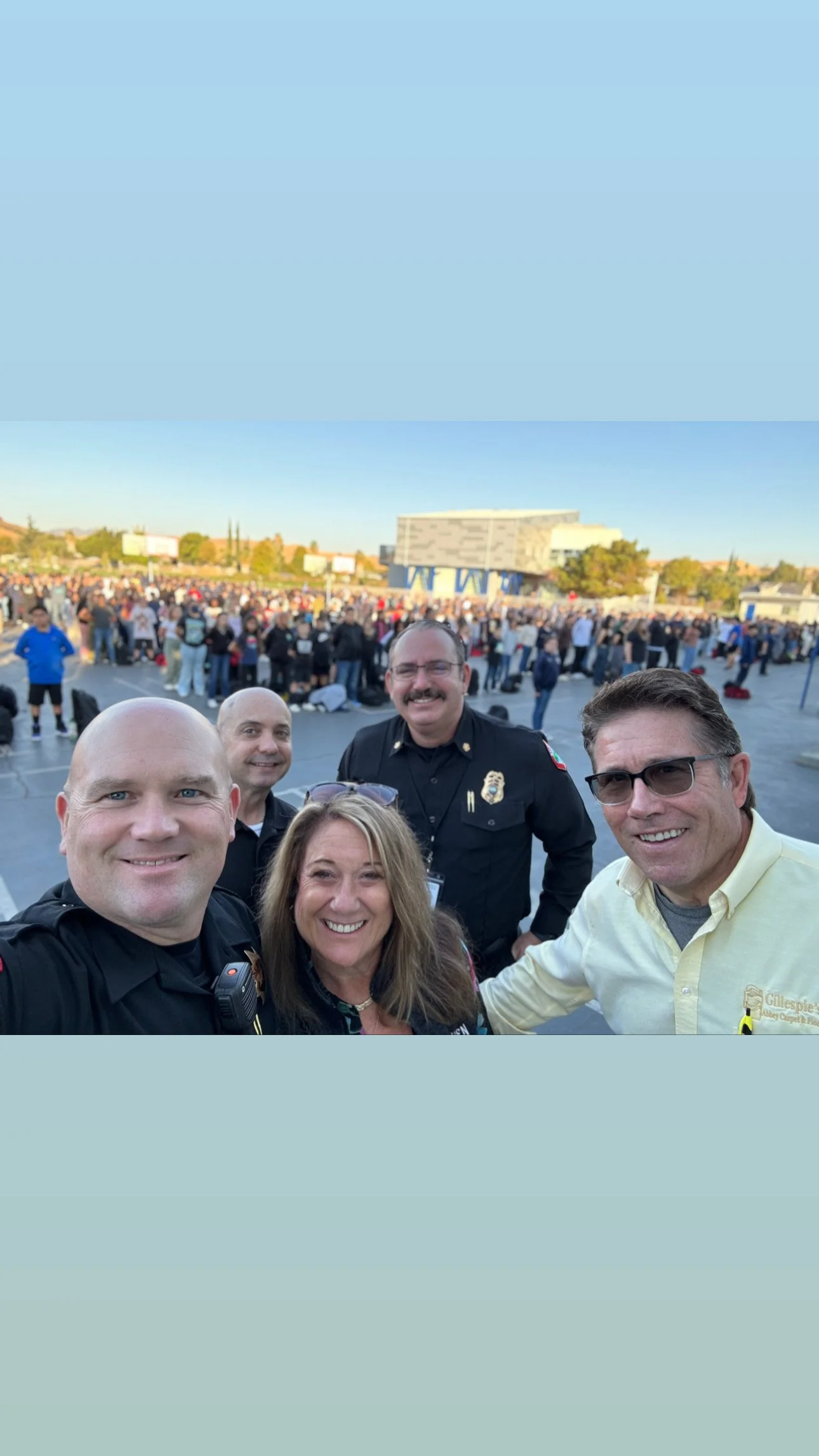 Group of five people taking a selfie outdoors at a large event, with a crowd of people in the background and a modern building.