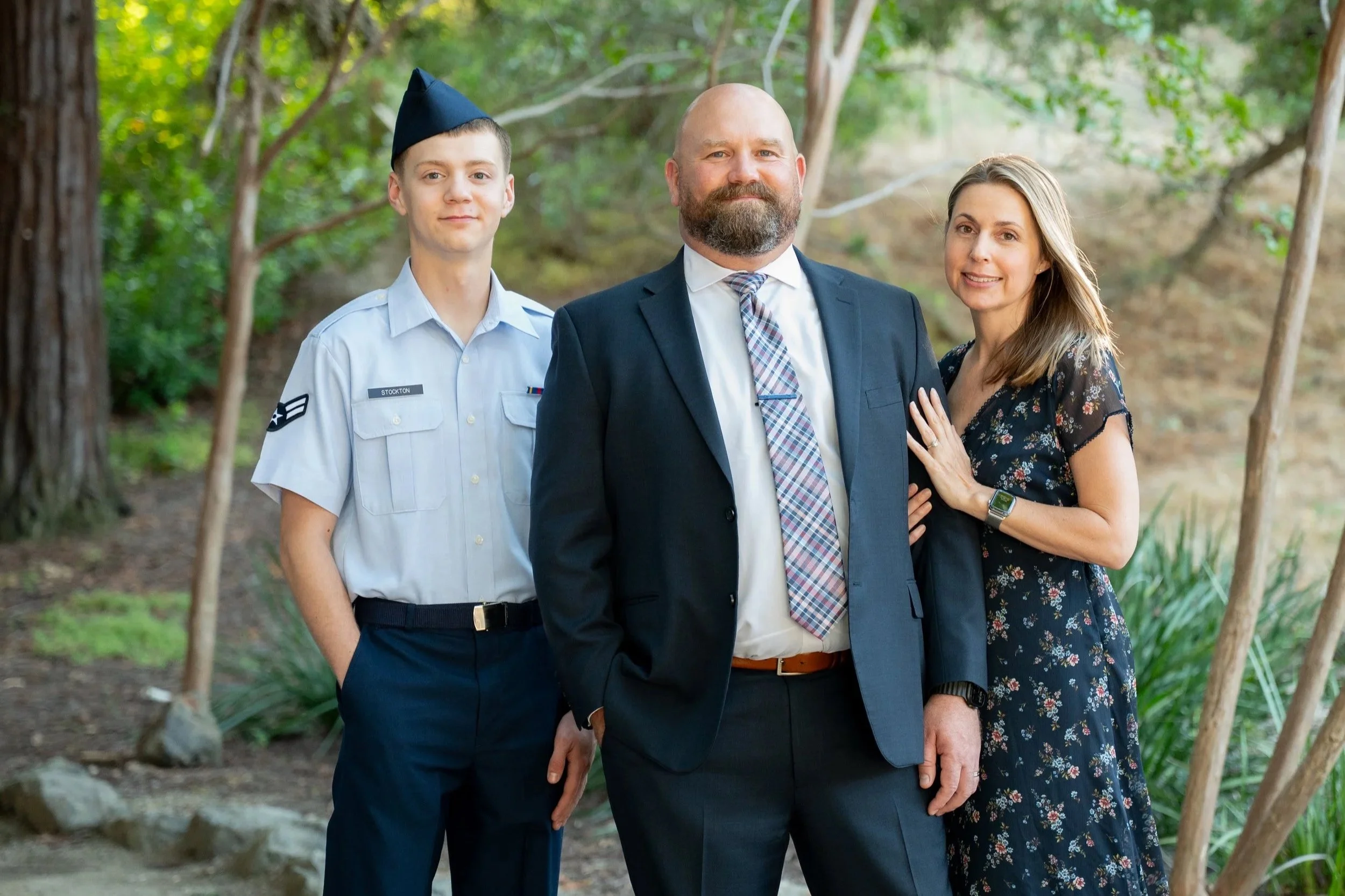 A family of three standing outdoors in a lush green setting, all dressed formally with the man in a suit, the woman in a floral dress, and the young man in a light blue shirt and military uniform.