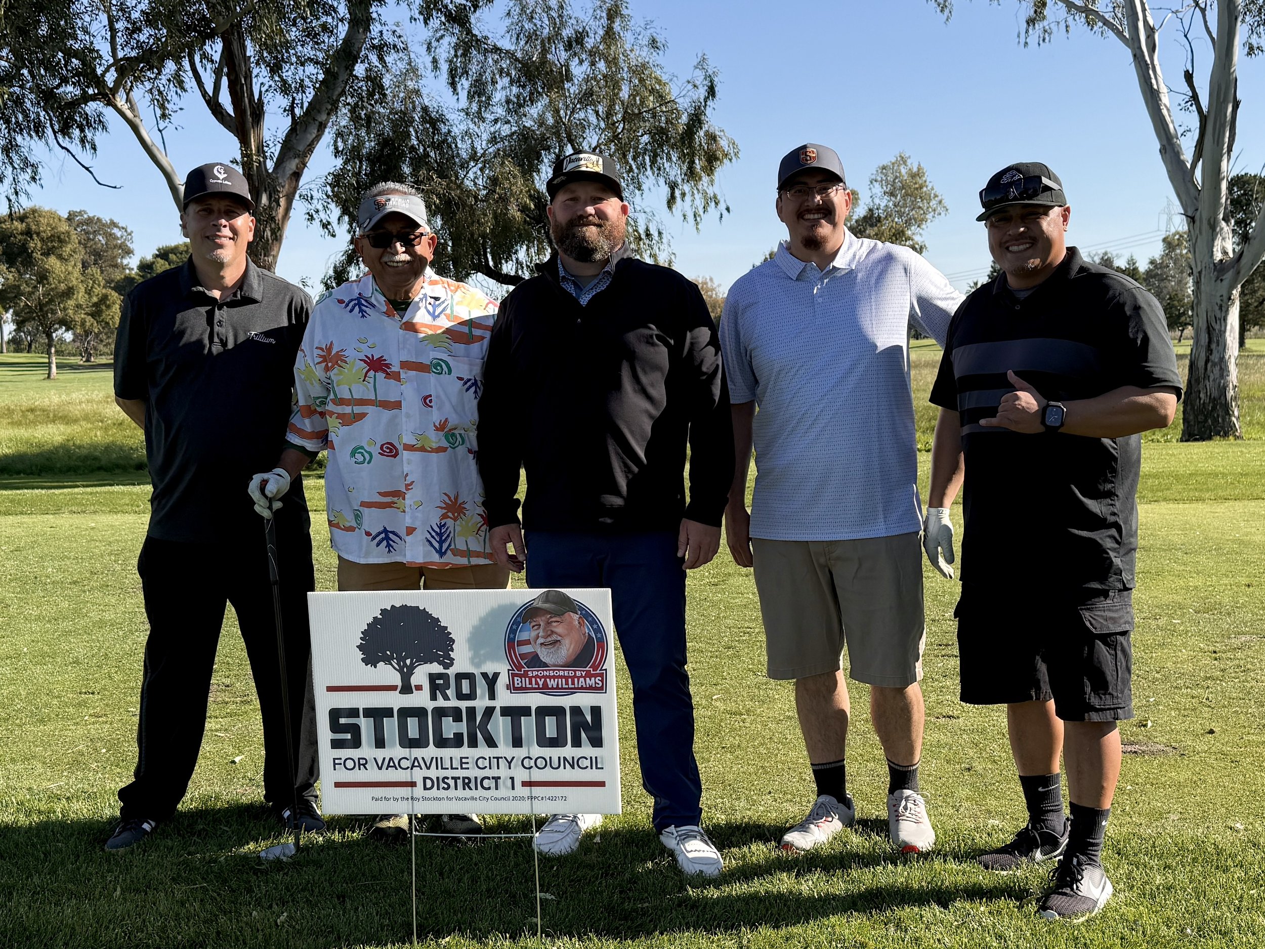 A group of five men standing on a golf course, holding a political campaign sign for Roy Stockton running for Vacaville City Council District 1. They are smiling and dressed casually, with trees and a clear blue sky in the background.