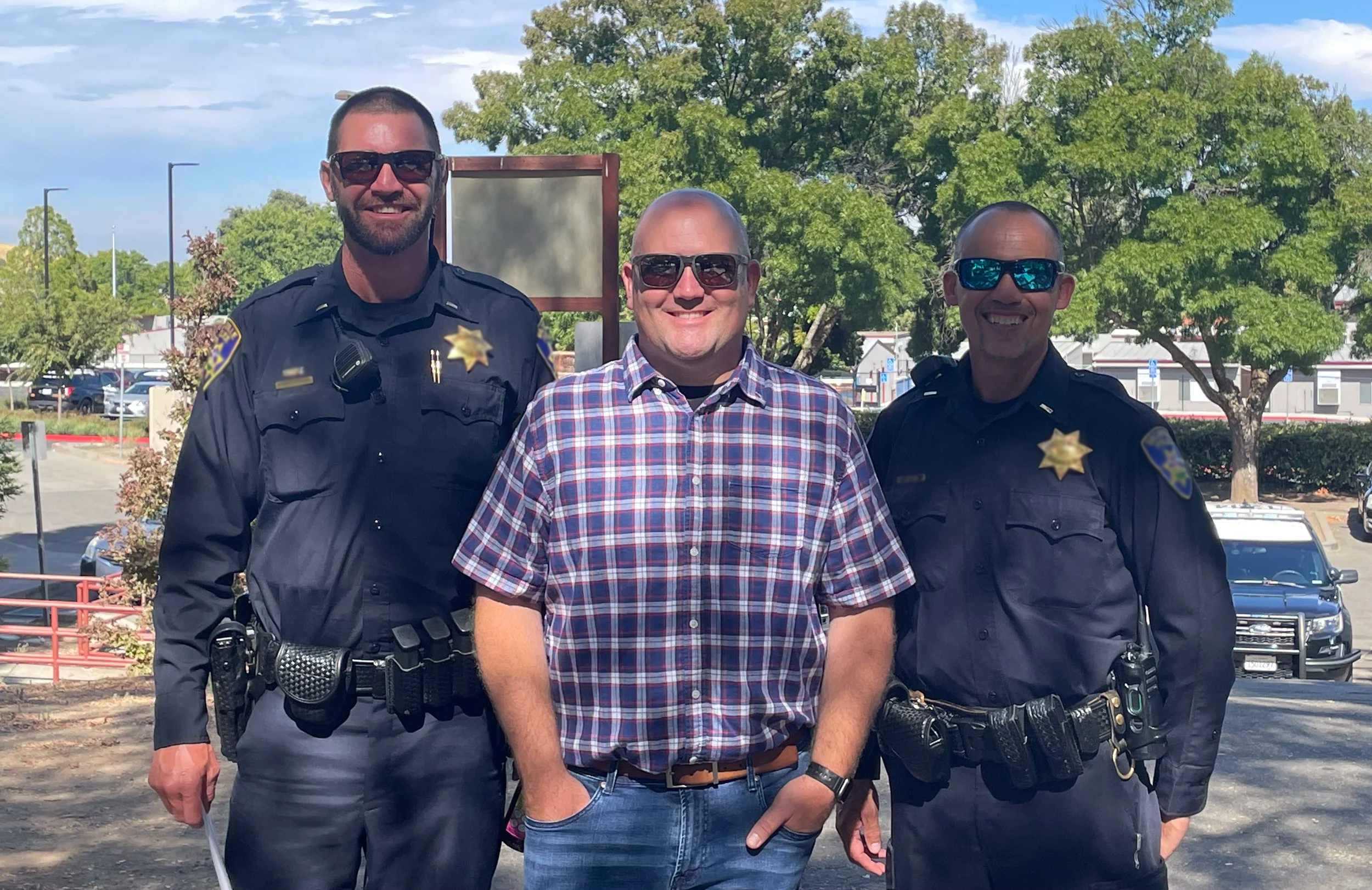 Three men, two in police uniforms and one in a plaid shirt, standing outdoors with trees and cars in the background