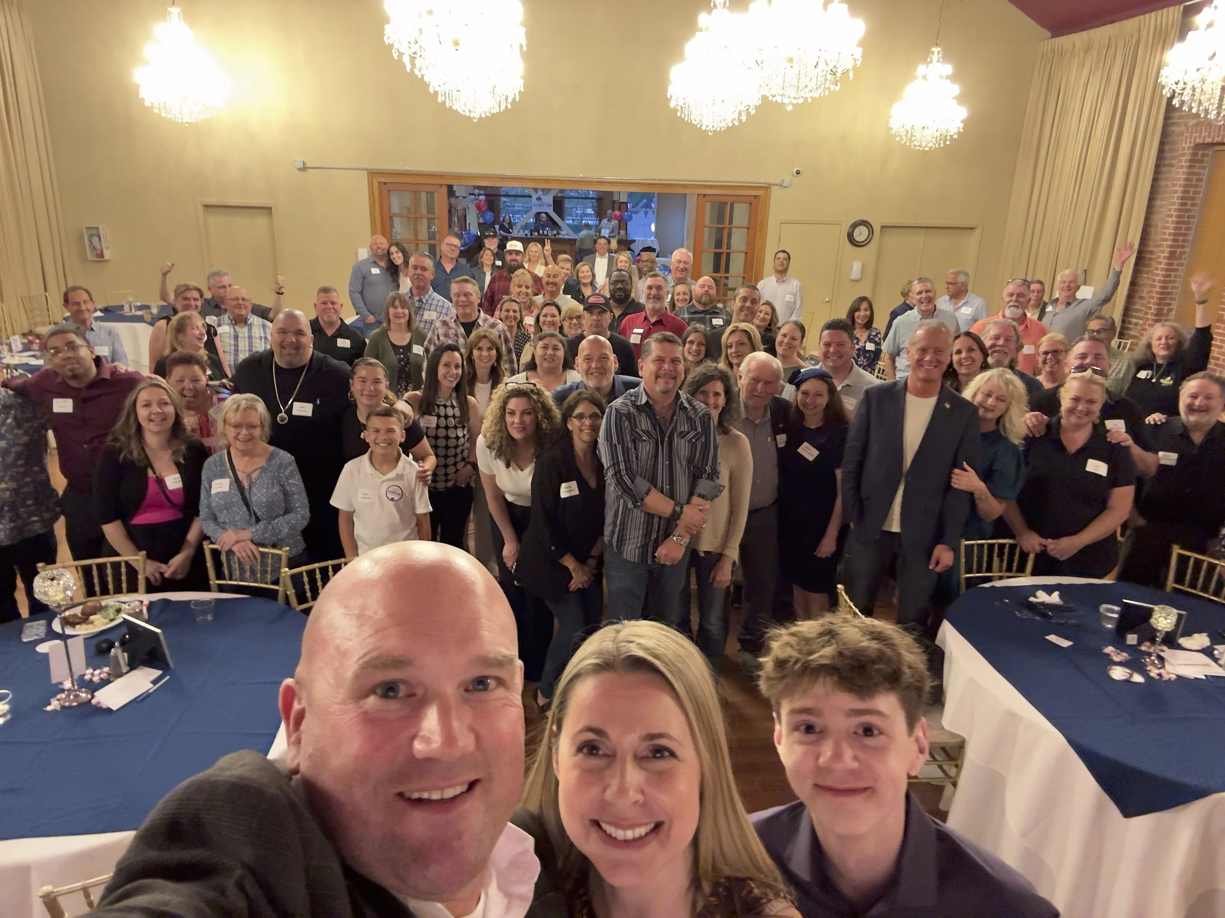 A large group of people gathered in a banquet hall for a celebration or event, smiling and posing for a group photo, with tables set with food and drinks in front of them.