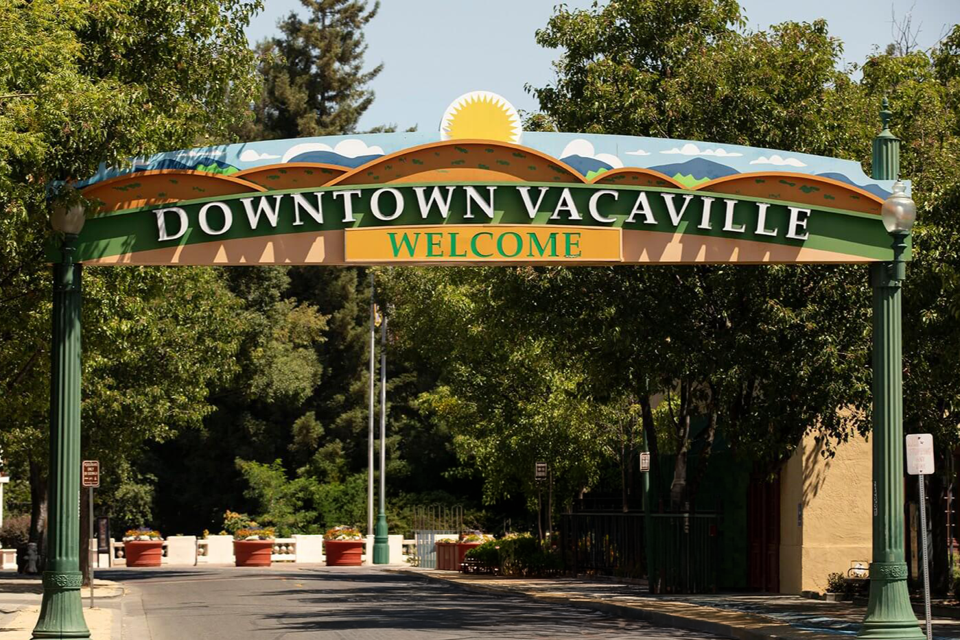 Sign that reads 'Downtown Vacaville Welcome' with a scenic background of mountains and a bright sun.
