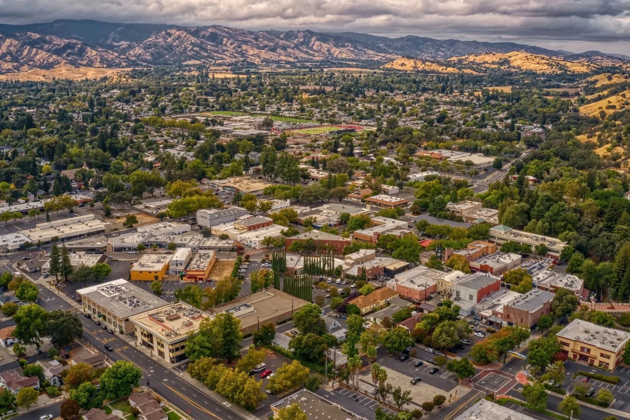 Aerial view of a city surrounded by trees with mountains in the background and cloudy skies overhead.