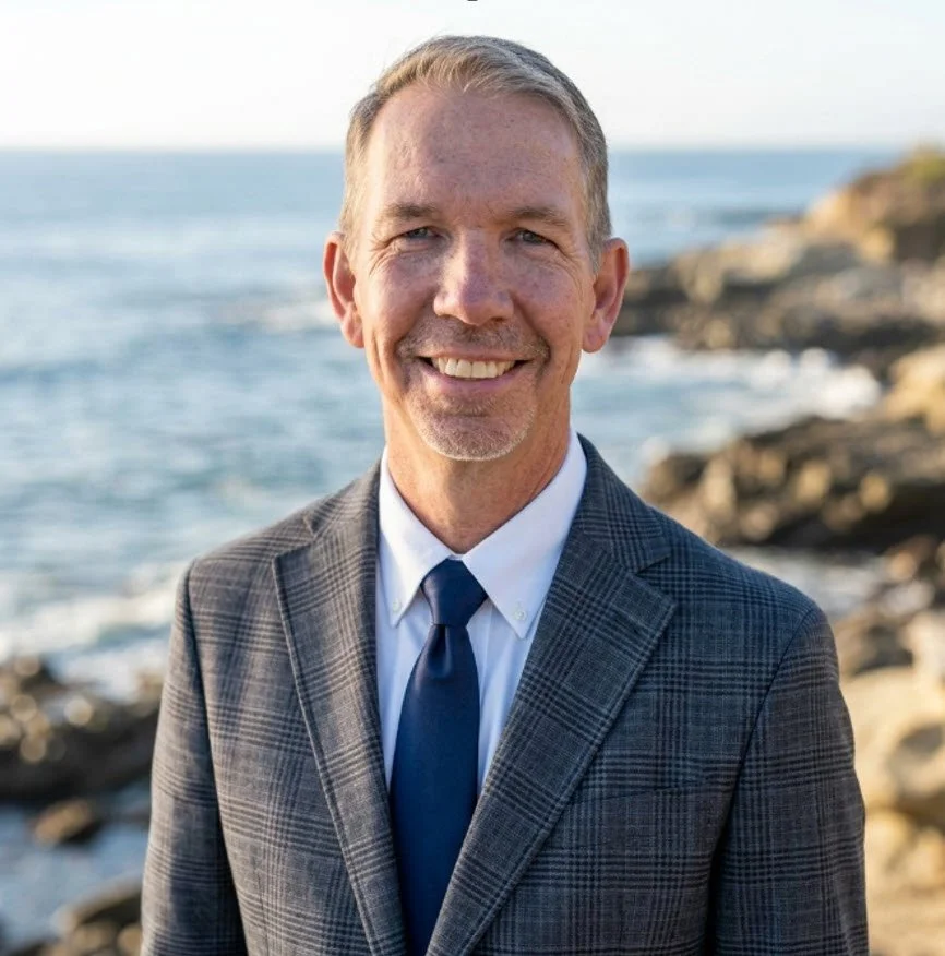 A smiling man in a suit standing by the rocky shoreline with the ocean in the background.