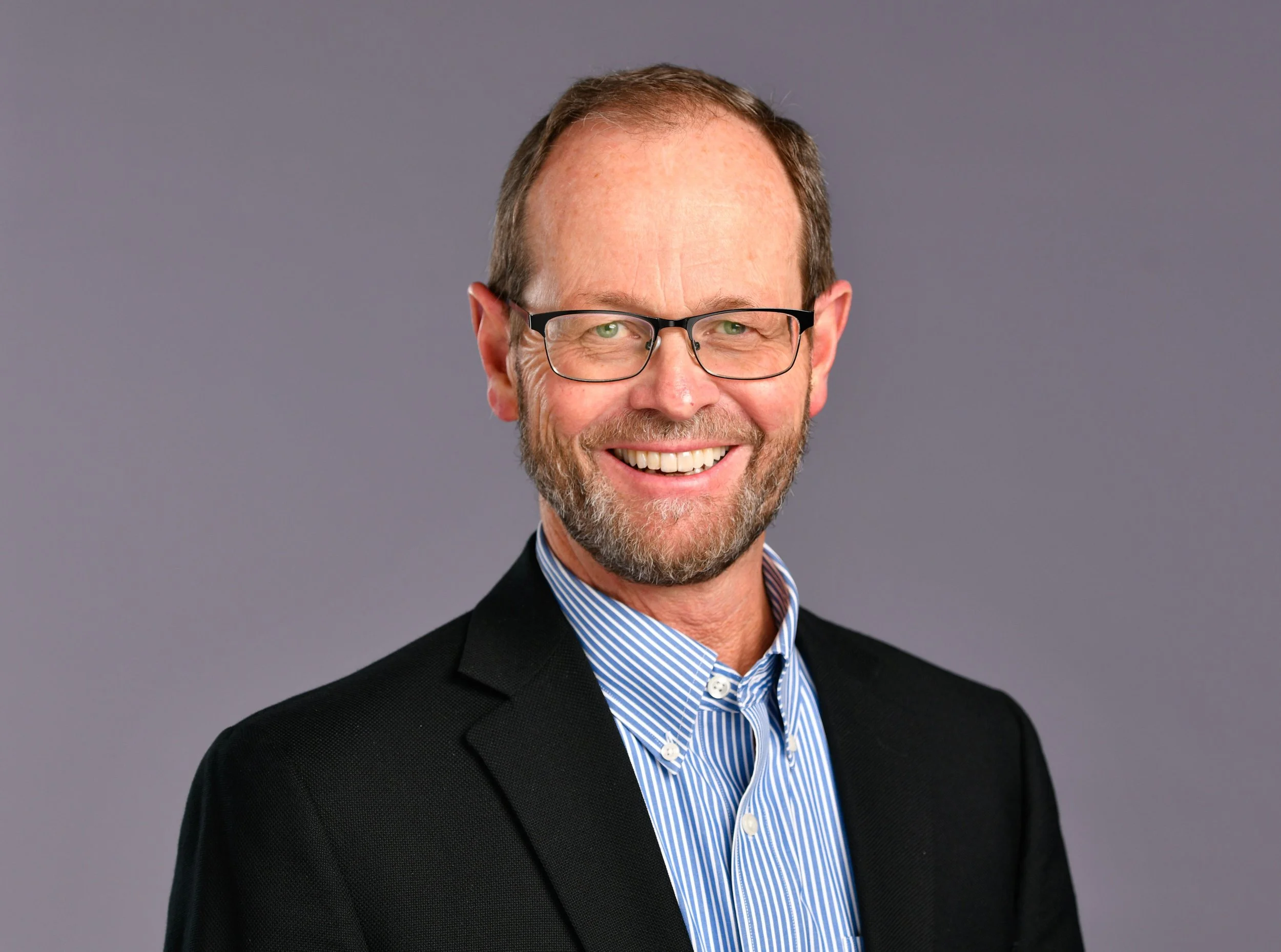 Portrait of a smiling middle-aged man with glasses, wearing a black blazer and a blue striped shirt, against a plain gray background.