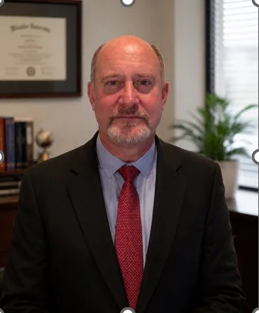 A middle-aged man with a beard and bald head, wearing a black suit, light blue shirt, and red tie, sitting in an office with a framed diploma and bookshelf in the background.