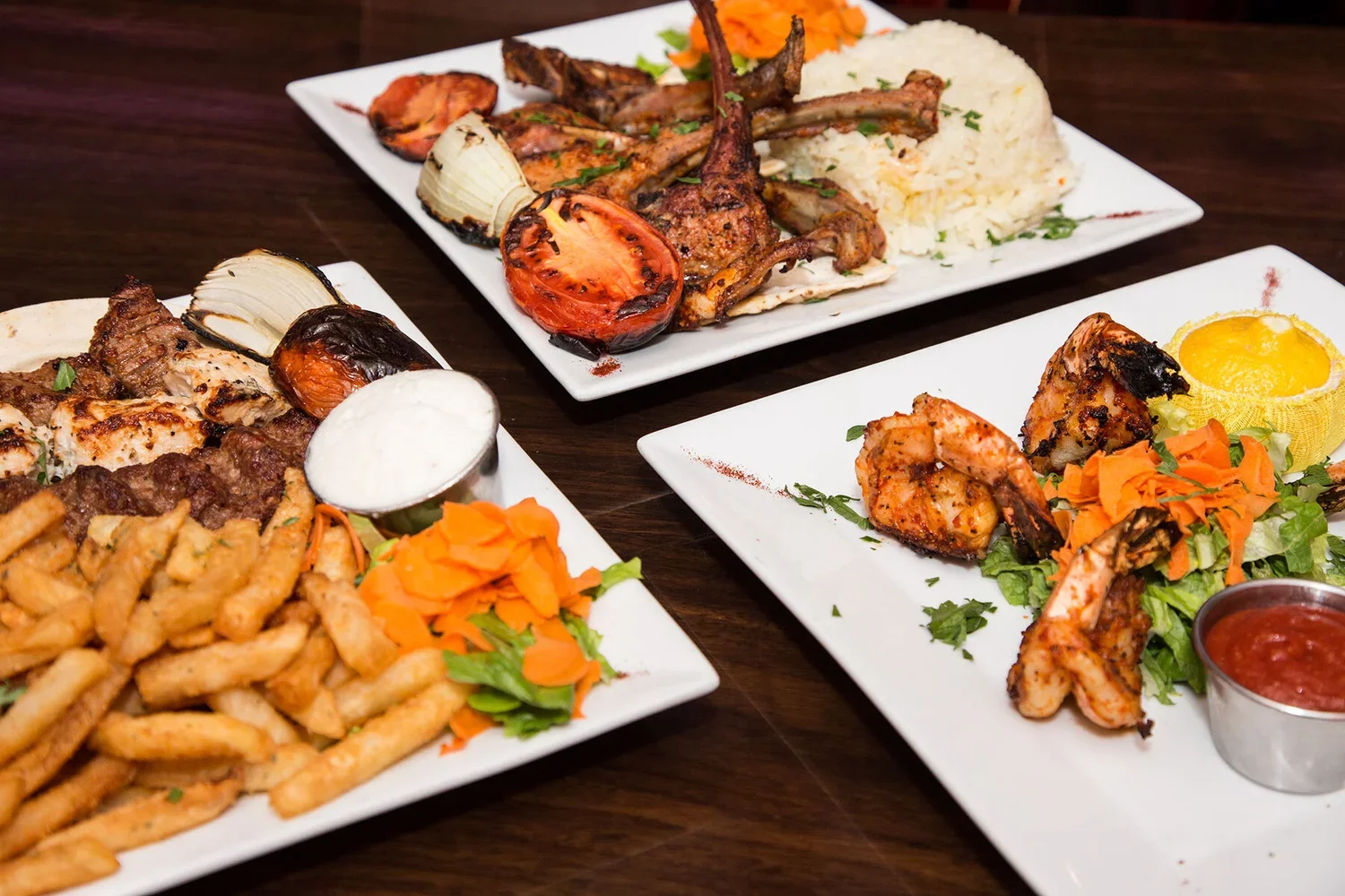 Three plates of grilled meat, vegetables, rice, and French fries on a wooden table