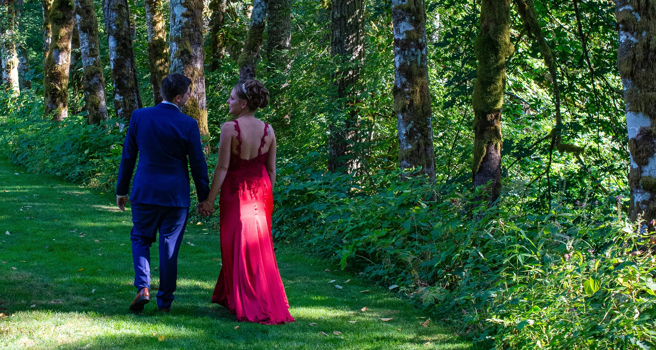 A couple dressed in formal attire walking hand in hand through a lush green forest. The woman is wearing a long, elegant red dress, and the man is in a dark suit. Sunlight filters through the trees, creating a serene and romantic atmosphere.