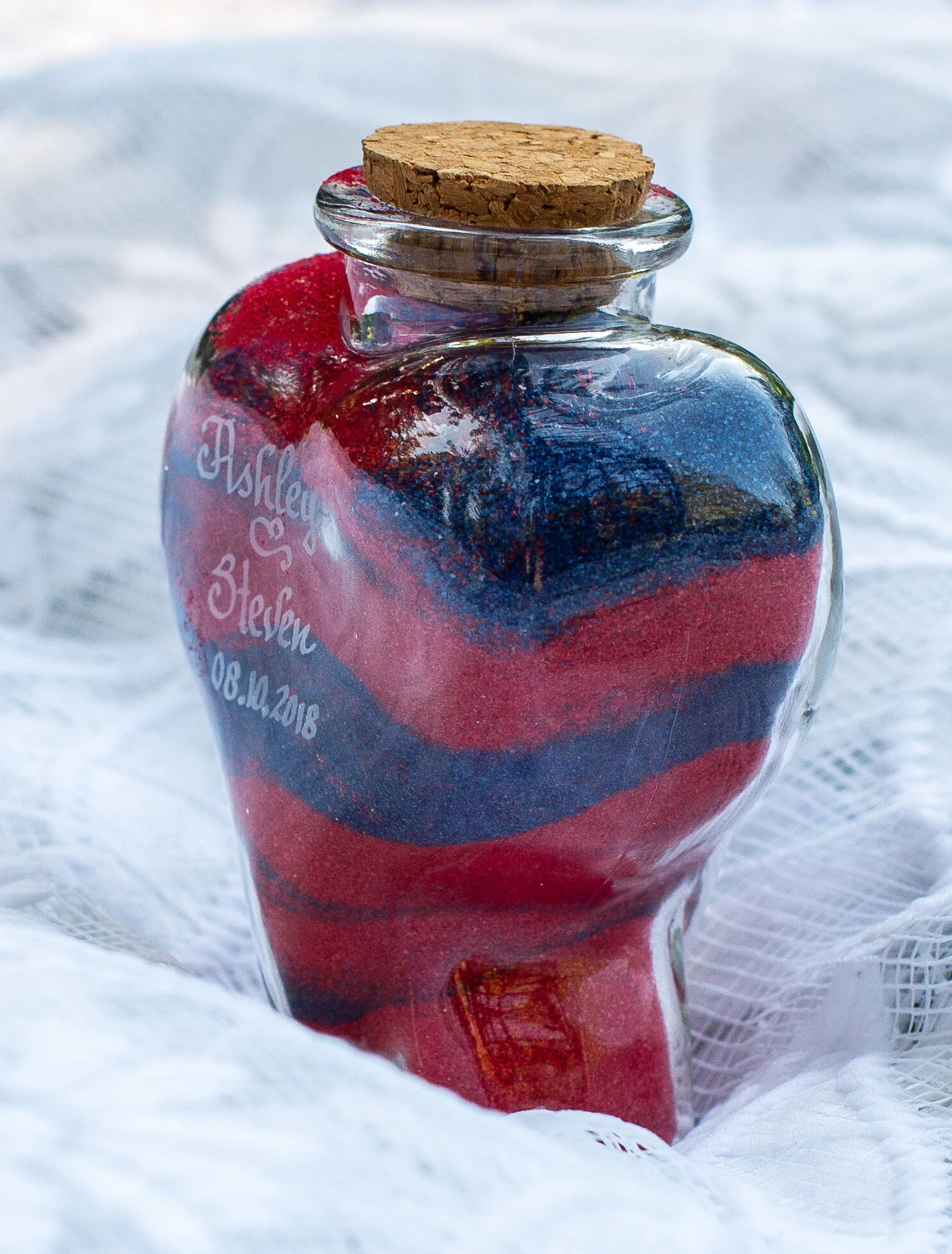 A glass jar with a cork lid containing red, blue, and black colored sand, placed on a white fabric surface.