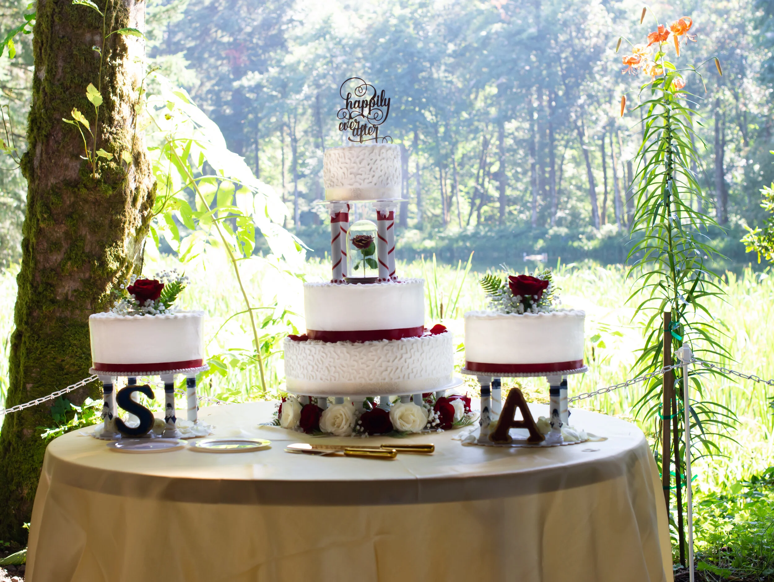 Wedding cake on a table with other smaller cakes, decorated with red and white roses, set outdoors in a forested area.