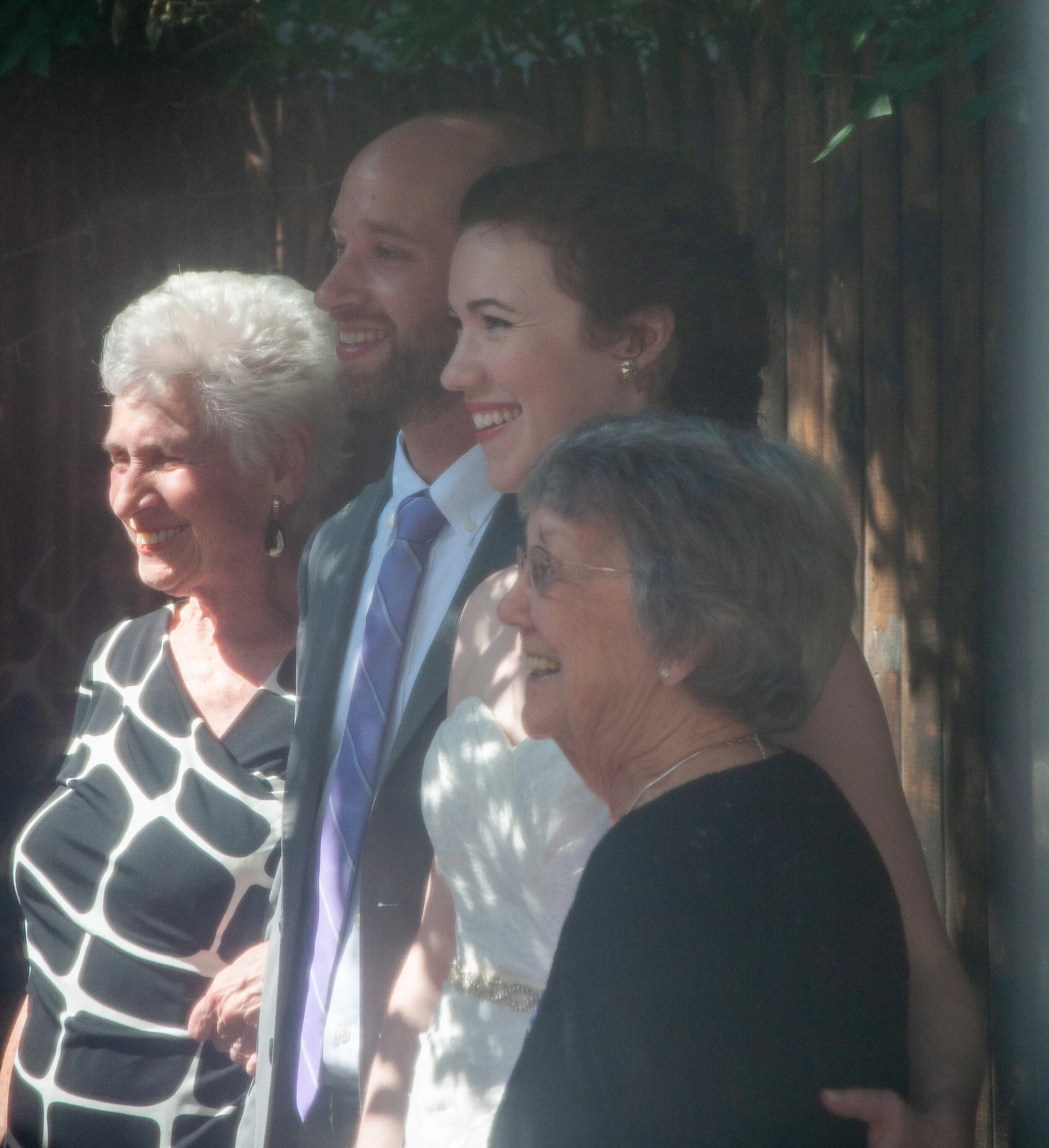 Four people standing together, smiling, outdoors near a wooden fence, with sunlight filtering through trees.