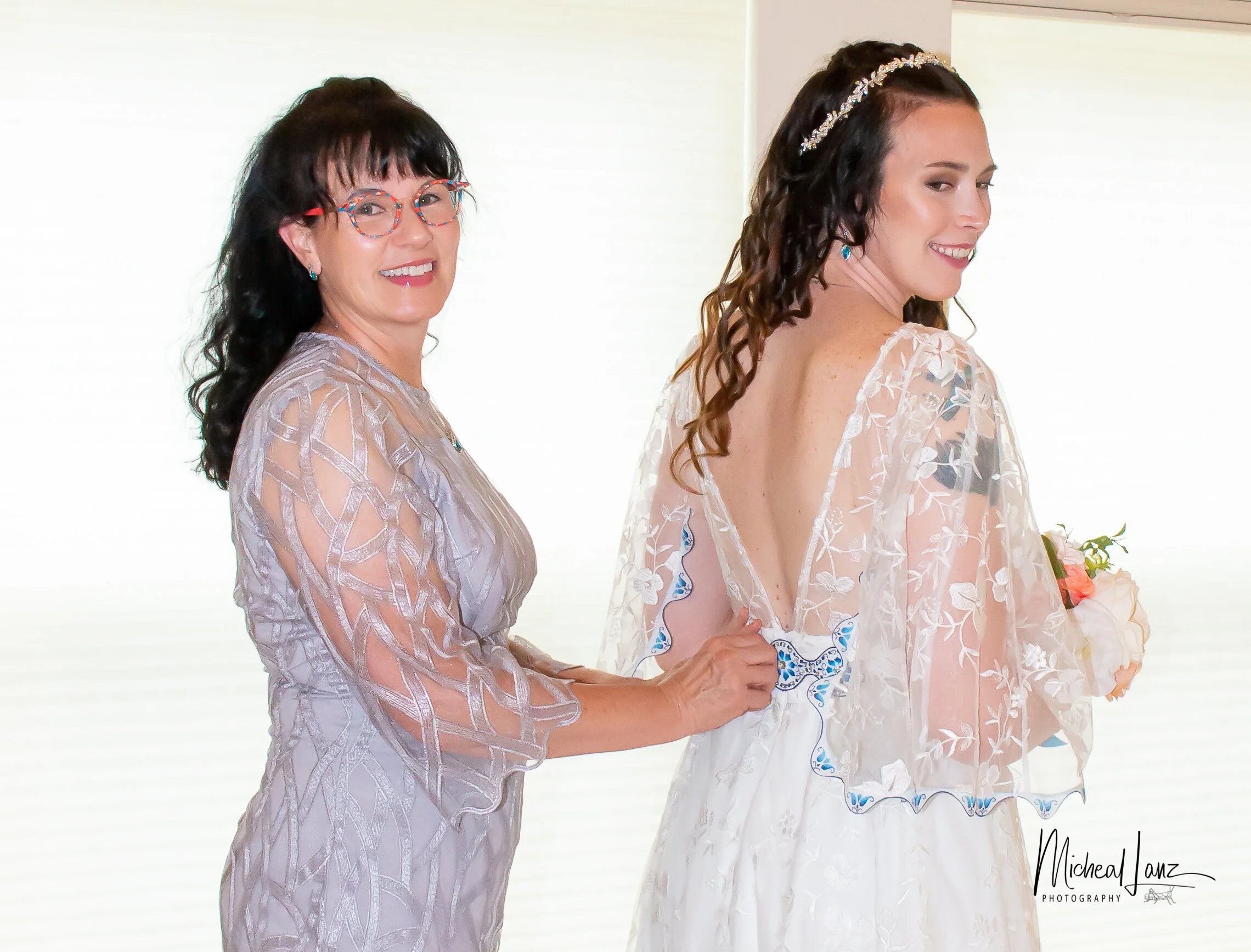A woman in a floral lace dress with dark, curly hair and a headband, smiling and holding hands with an older woman with glasses, dark hair, and a patterned dress, in a bright indoor setting.
