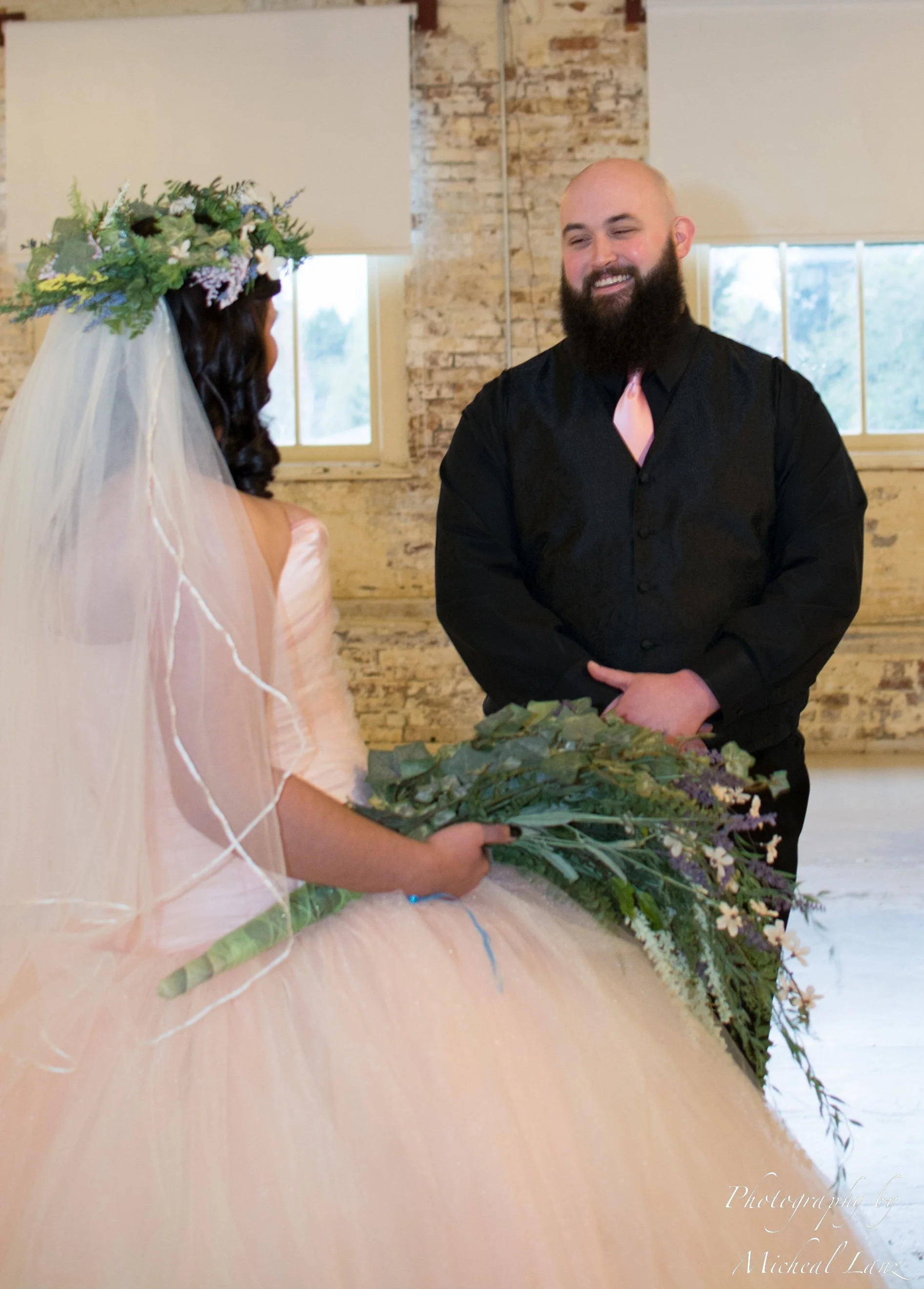 A woman with a floral crown and veil holding a bouquet of flowers facing a man with a beard, dressed in black with a pink tie, smiling during a wedding ceremony in an indoor venue with brick walls and windows in the background.