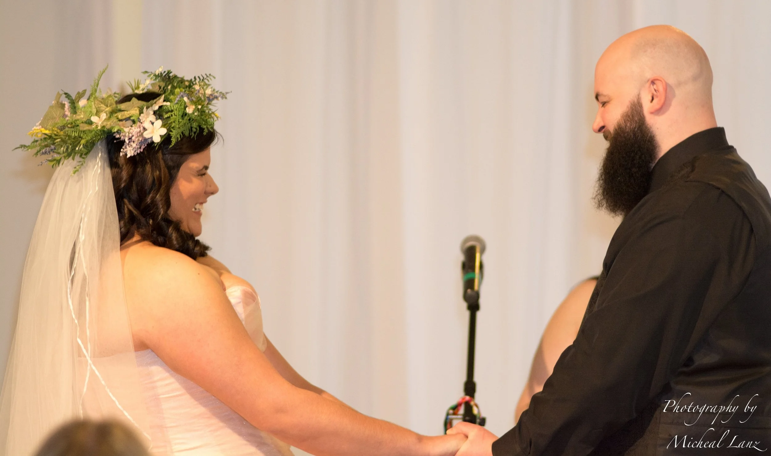 A bride with dark hair, wearing a floral crown and veil, holding hands with a bald groom with a beard, during their wedding ceremony.
