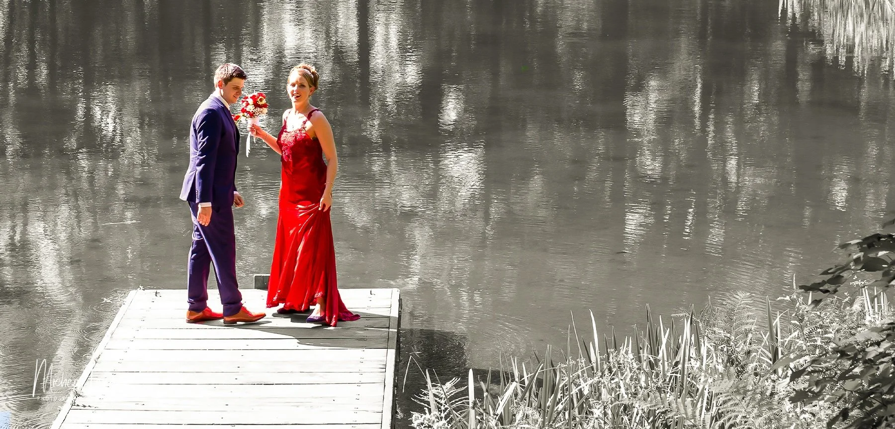 A couple stands on a wooden dock by a lake. The woman in a red dress holds a bouquet of flowers, and the man is in a navy suit. The background and surroundings are in black and white, while the couple is in color.