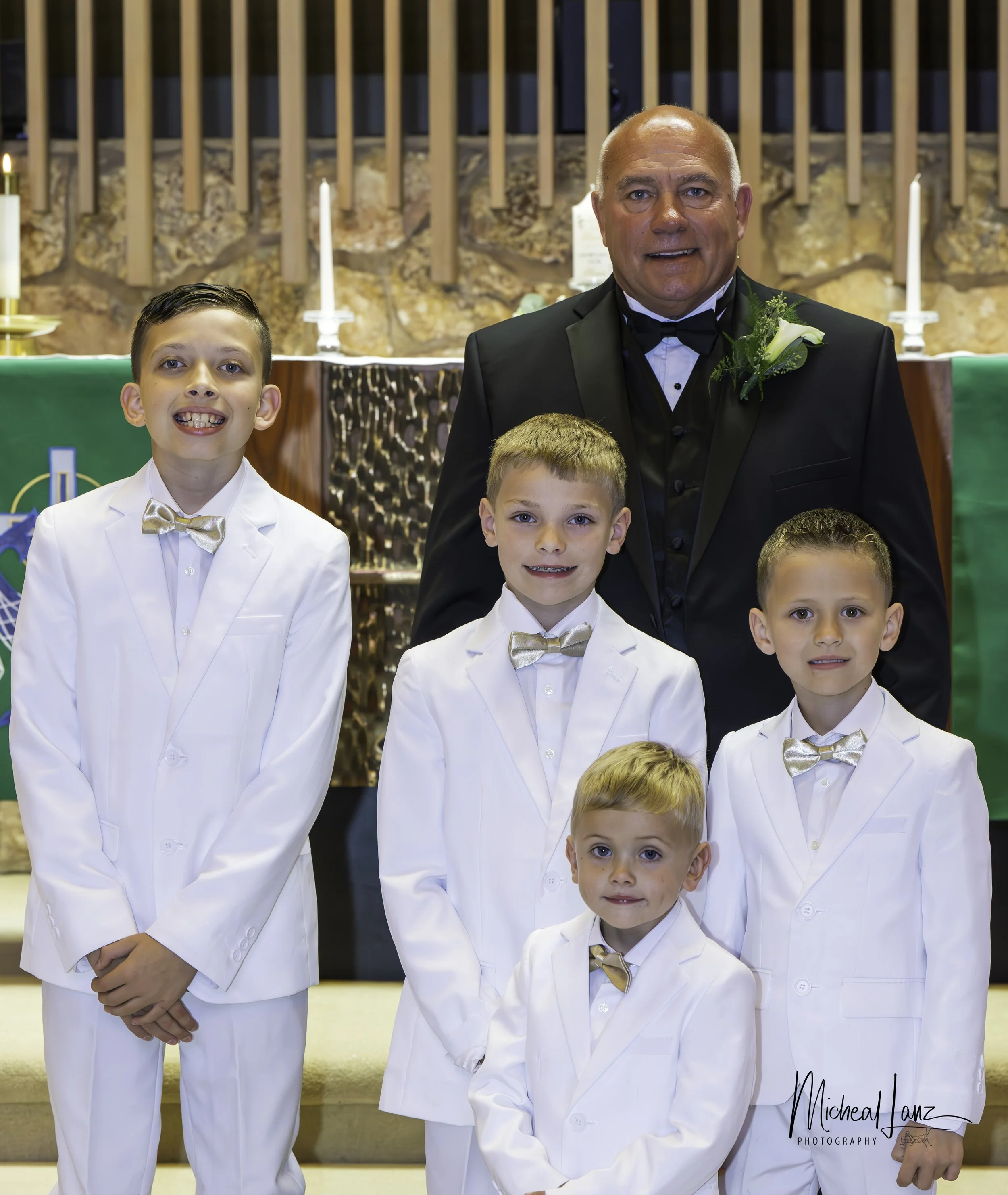 A group of five young boys dressed in white suits with gold bow ties, standing in front of an altar with lit candles, a green cloth, and a background of wooden beams and stone wall, during a religious ceremony.