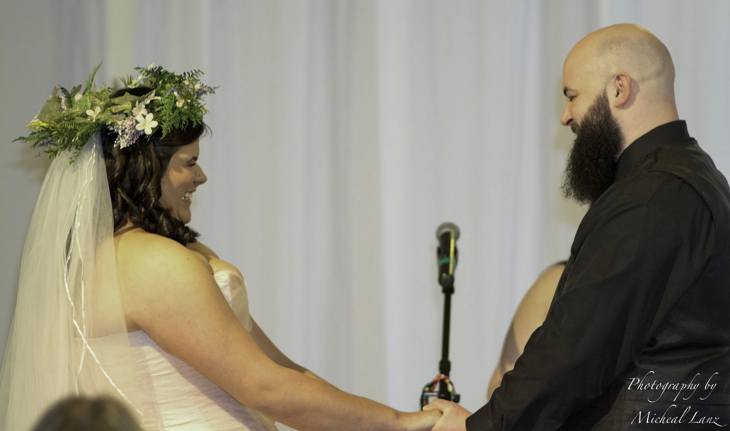 A woman with a flower crown and veil holding hands with a man in a black shirt during a wedding ceremony.