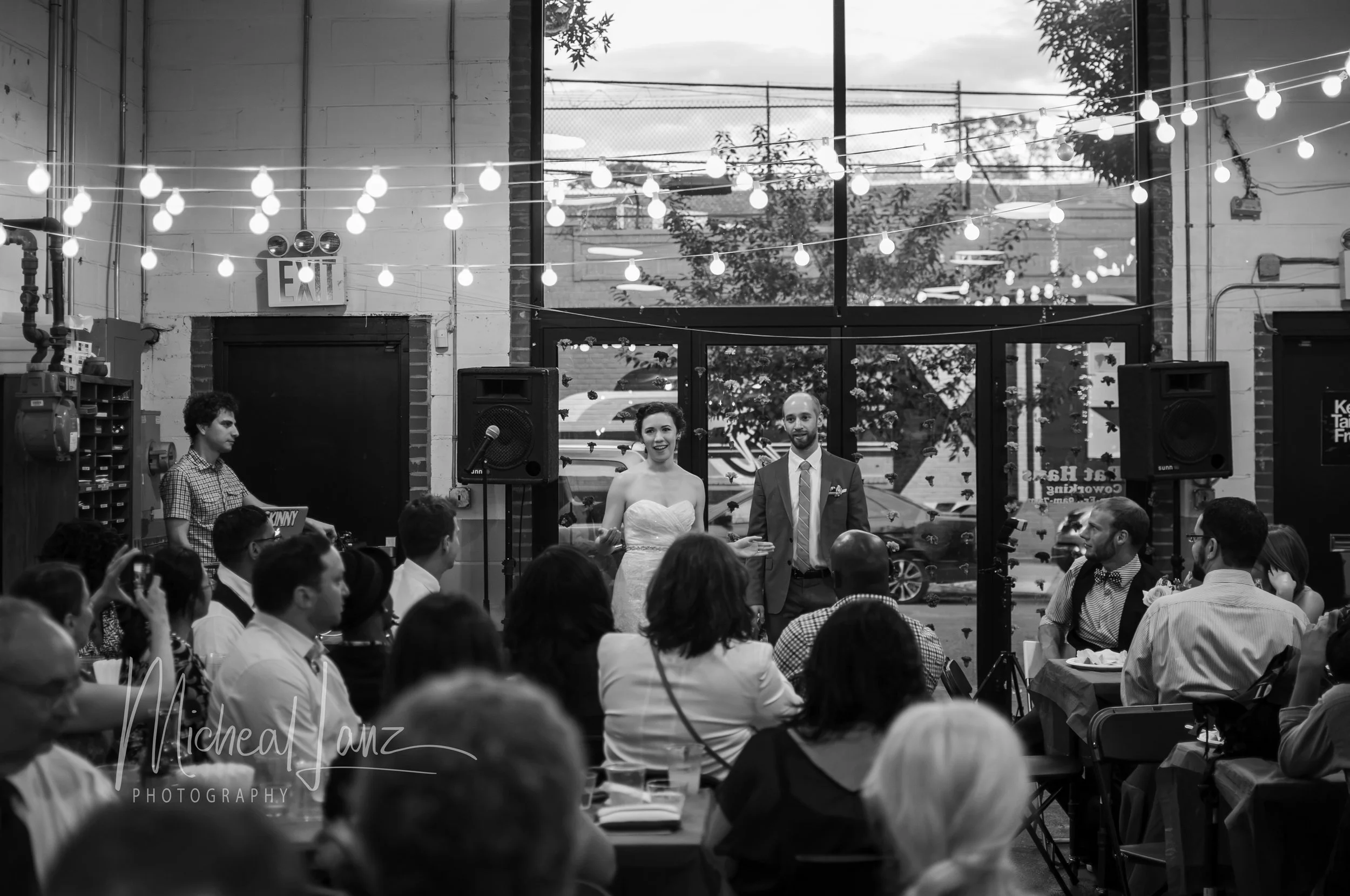 A black-and-white photo of a wedding ceremony inside a venue with large windows and string lights. The bride and groom stand in front of seated guests, with the bride wearing a strapless dress and the groom in a suit.